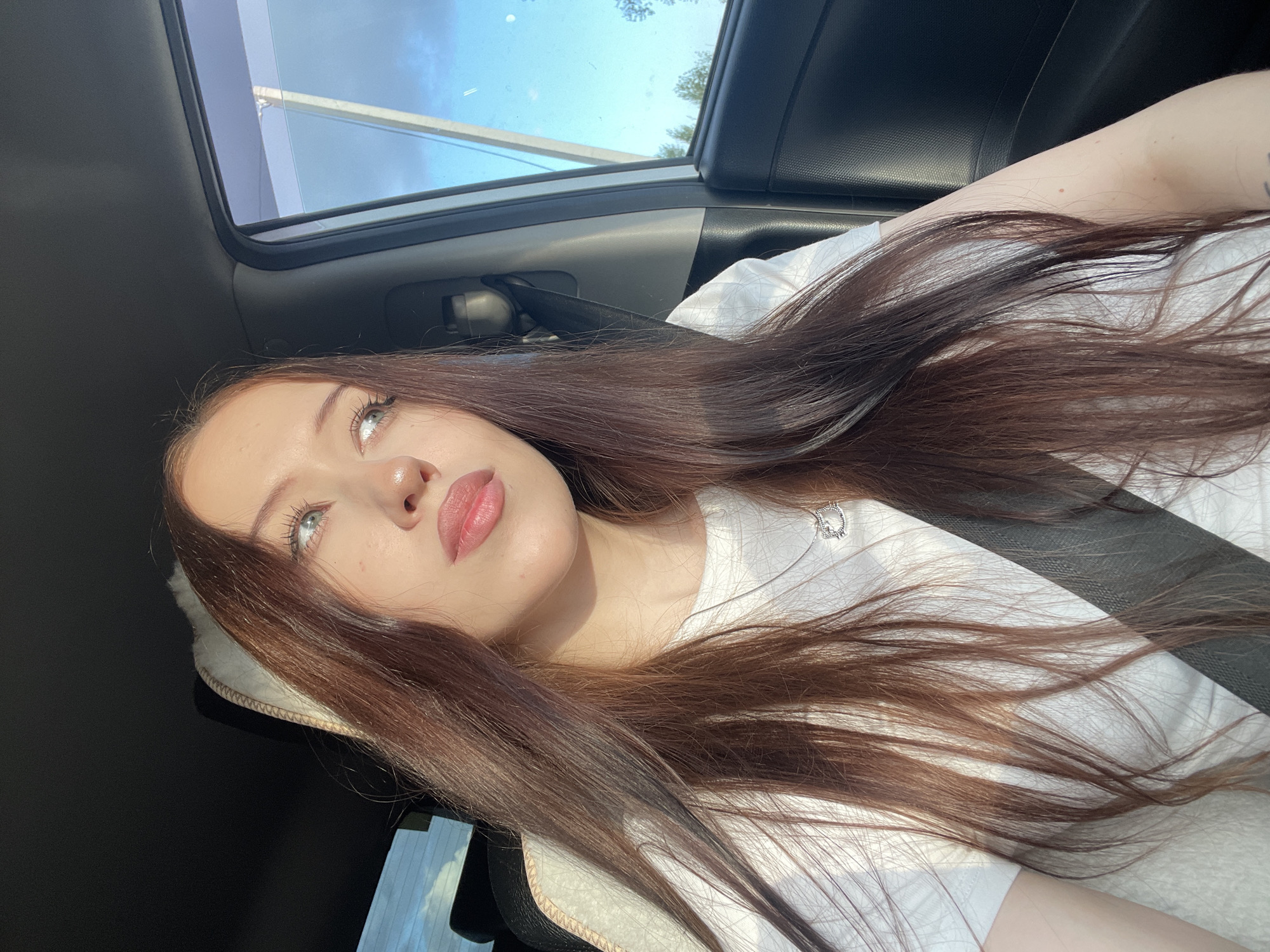 A young Caucasian woman with long, dark brown hair is seated in the passenger seat of a vehicle, looking out the window. She is wearing a white t-shirt and a silver necklace with a heart-shaped pendant. The interior of the car is visible, including the seatbelt, car door, and a portion of the dashboard. Sunlight streams in from the right side of the frame, illuminating her face and hair. The window shows a view of the sky with scattered clouds and some distant trees, suggesting an outdoor location, possibly on the outskirts of a city like Gavrilovka, Russia.