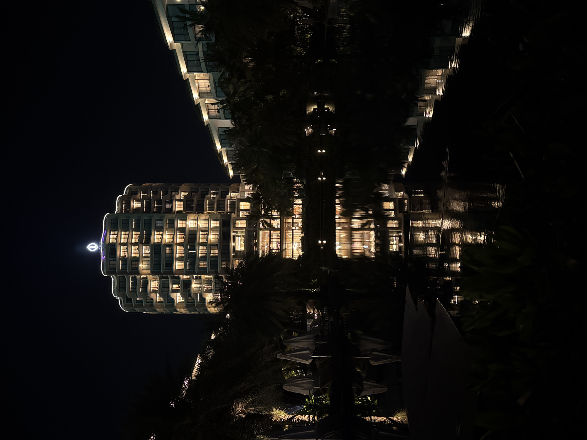 The image shows a nighttime scene in Phú Quốc, Vietnam. Two illuminated buildings stand with their reflections visible in a body of water, likely a pool or a calm part of the sea. The buildings, with multiple stories and illuminated windows, exhibit modern architecture. Palm trees and other foliage are present, adding to the tropical atmosphere. In the foreground, dark shapes suggest umbrellas and other outdoor furniture or structures.