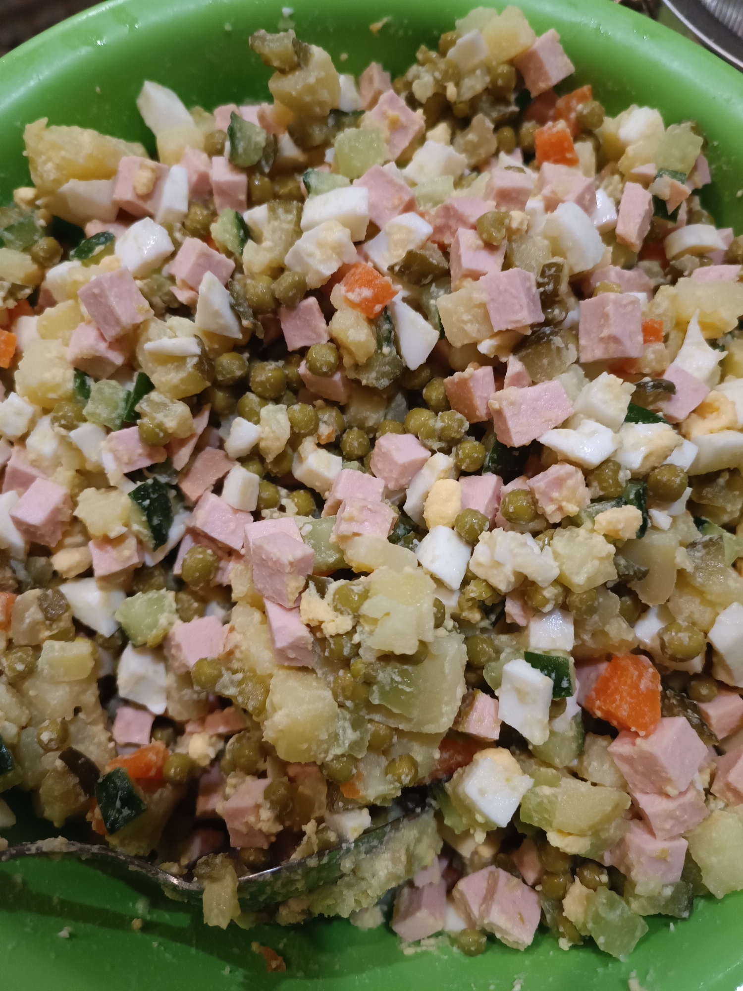 The image shows a close-up view of a green bowl filled with a salad. The salad appears to be a potato-based salad with diced ingredients including ham, hard-boiled eggs, peas, pickles, and carrots. A spoon is partially visible in the lower left corner, suggesting the salad is ready to be served or eaten. The background is out of focus, but the green color of the bowl is prominent. The scene is indoors, and there is no indication of the specific location within Zaporizhzhia, Ukraine. There are no people visible in the image.
