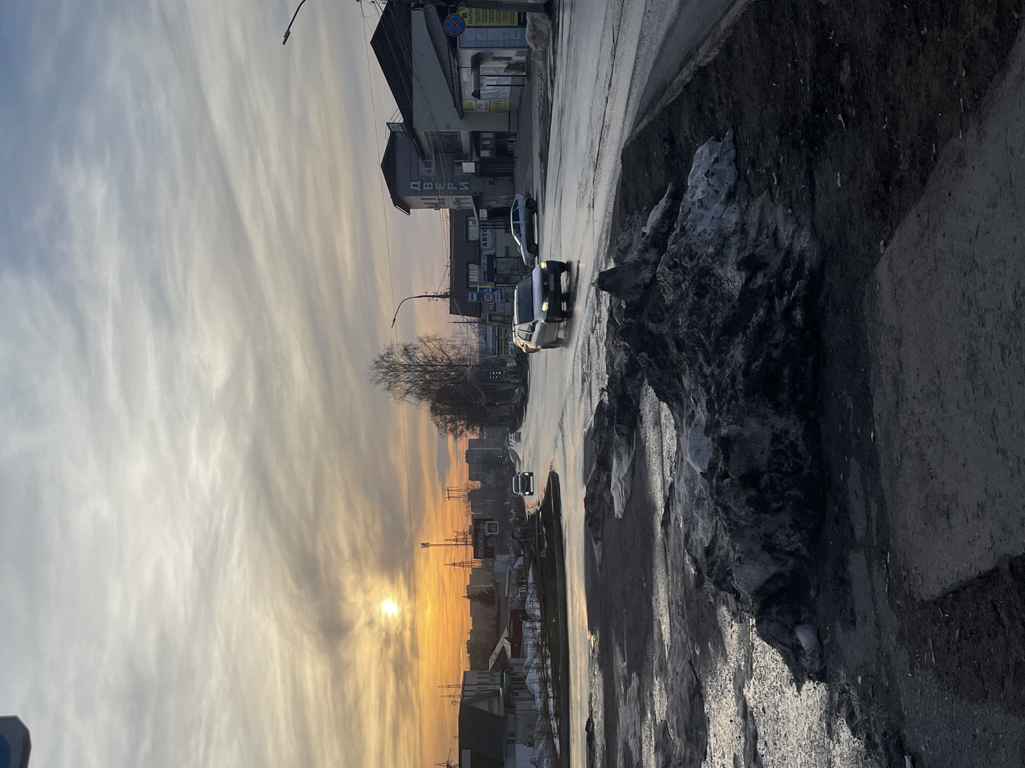 The image depicts an urban street scene in Chaikovsky, Russia, captured during a sunset or sunrise. The sky is characterized by an orange glow from the low sun on the left horizon, blending into grey and white streaky clouds across the frame.
The foreground consists of a roadside area featuring large, dark, dirty piles of melting snow and exposed patches of wet, muddy ground. The asphalt roadway is wet and reflective, indicating the presence of water or slush.
Multiple vehicles are present on the road. A silver sedan with its headlights illuminated is prominently visible in the mid-ground, traveling away from the viewer on the right side of the road. Other vehicles are discernible further down the street in both directions.
To the right of the road, several multi-story commercial buildings are visible. One prominent building features the Cyrillic word "ДВЕРИ" (DOORS) written vertically on its facade. An adjacent building displays a sign with the word "КОЛБАСА" (SAUSAGE) and a red circular traffic sign. On the left side of the street, lower-profile buildings, utility poles, and what appear to be industrial structures or power line towers are silhouetted against the bright horizon. A yellow pedestrian crossing sign and a larger sign with "Реклама и Вывески" (Advertising and Signs) are also visible on the left. Bare deciduous trees line parts of the street, consistent with a late winter or early spring setting.