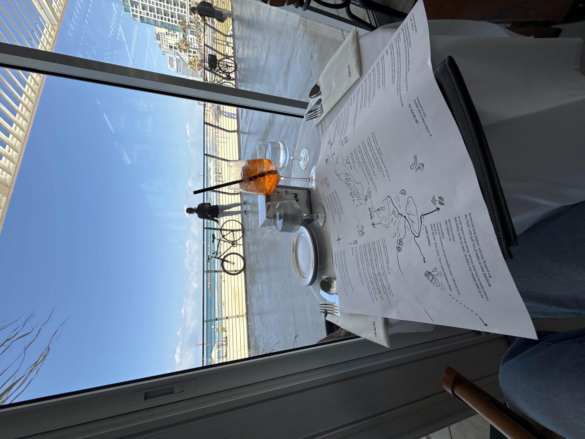 The image presents a scene from a restaurant in Tel Aviv, Israel, overlooking the beach and Mediterranean Sea. Through the large window, people can be seen walking and biking along the promenade. In the restaurant, a table is set with a glass of Aperol spritz and other glasses, a plate, cutlery, and a folded napkin. A menu, partially visible, shows Hebrew text listing various food items and descriptions. The menu is placed on top of a black wallet or folder. A person is sitting at the table, partially visible on the right side of the frame.