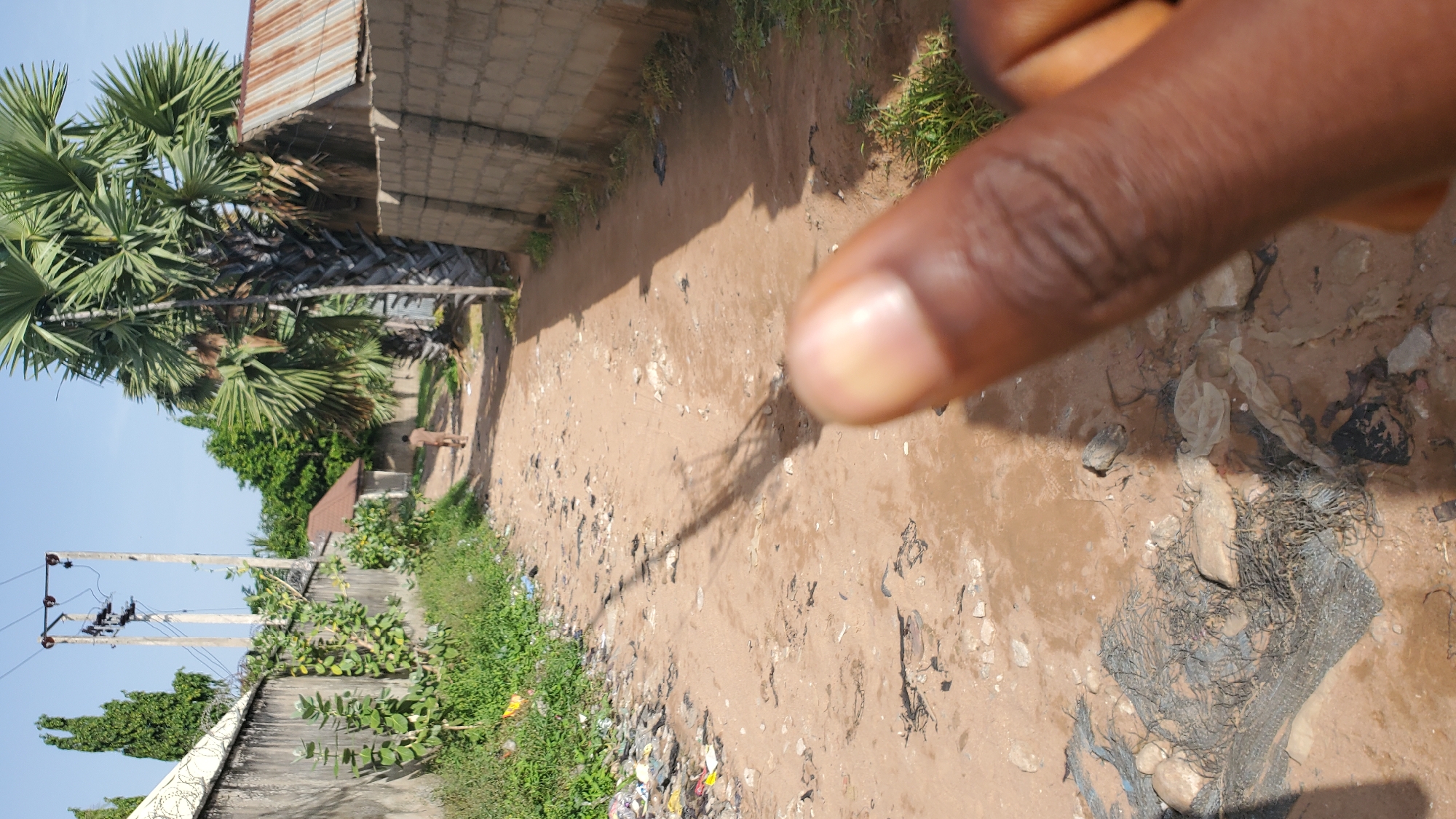 The image depicts a dirt road in a residential area of Jalingo, Nigeria. The road is lined with walls and vegetation, and there is debris scattered on the ground. In the foreground, a blurred human thumb is visible. A person is walking away in the distance down the road. Tall palm trees and power lines are also present in the scene, suggesting a somewhat rural or underdeveloped urban environment. The sky is clear and blue.