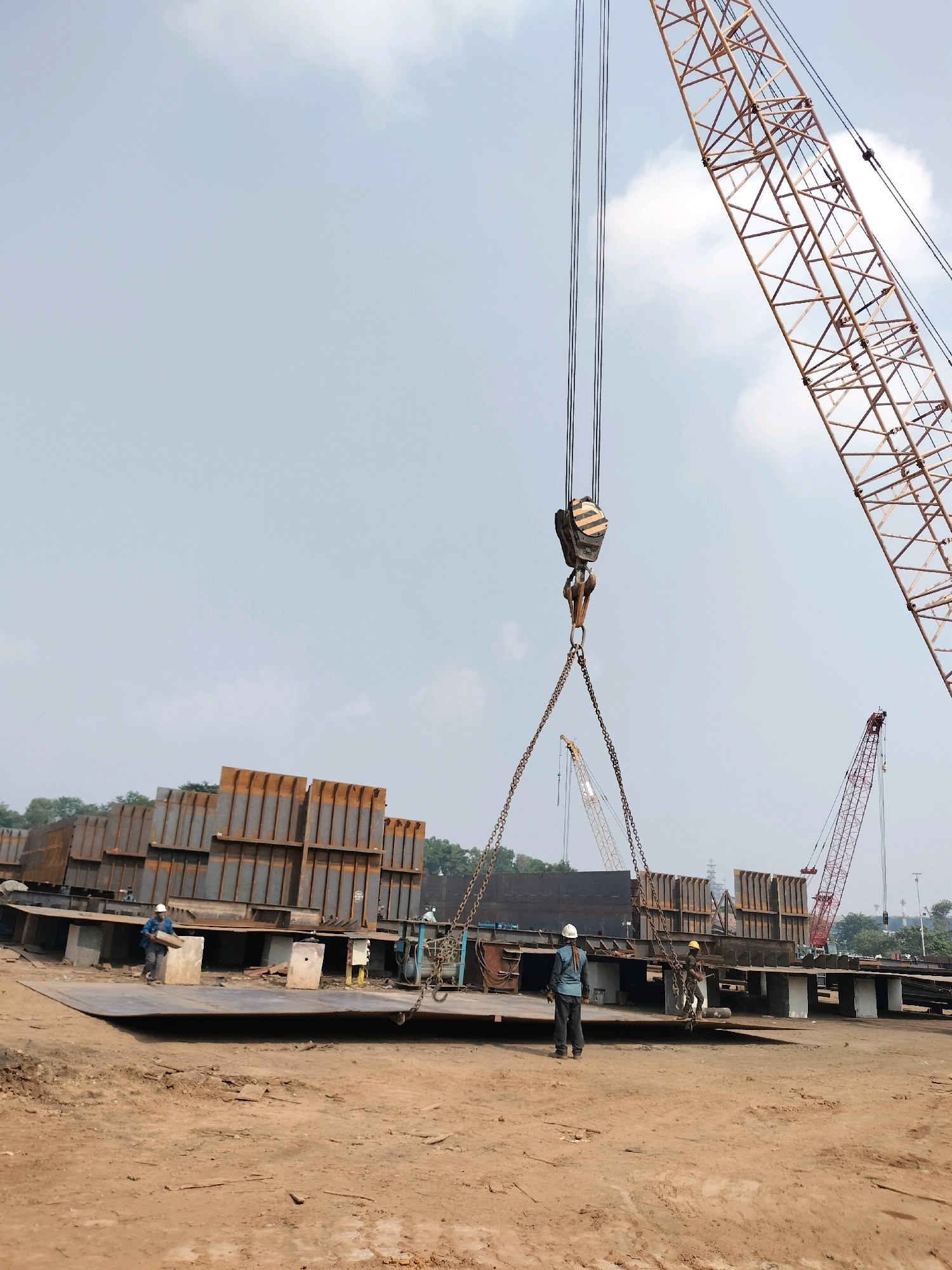 This is a daytime, eye-level, outdoor photograph taken at a shipyard in Batam, Indonesia. The image features a construction site with large steel structures and cranes. In the foreground, a large flat steel plate is positioned on the ground, supported by concrete blocks. Two construction workers are present. One worker, wearing a blue shirt and white hard hat, stands on the steel plate, holding a small object. Another worker, wearing a blue shirt, dark pants, and a white hard hat, is further back on the plate, facing away from the camera.
In the mid-ground, a stack of vertically oriented, large, rust-colored steel sections is visible. Several smaller, horizontally laid steel plates are also present, some supported by concrete blocks.
The background showcases several industrial cranes. A large yellow lattice boom crane dominates the right side of the frame, its boom extending high into the sky. To the left of this, a smaller red lattice boom crane is visible. Another lighter-colored crane is positioned further in the background. The sky is overcast with some light clouds. Trees and vegetation can be seen behind the steel structures. The ground appears to be a mixture of dirt and debris, typical of a construction or industrial environment. The overall scene suggests ongoing heavy industrial construction or fabrication work.