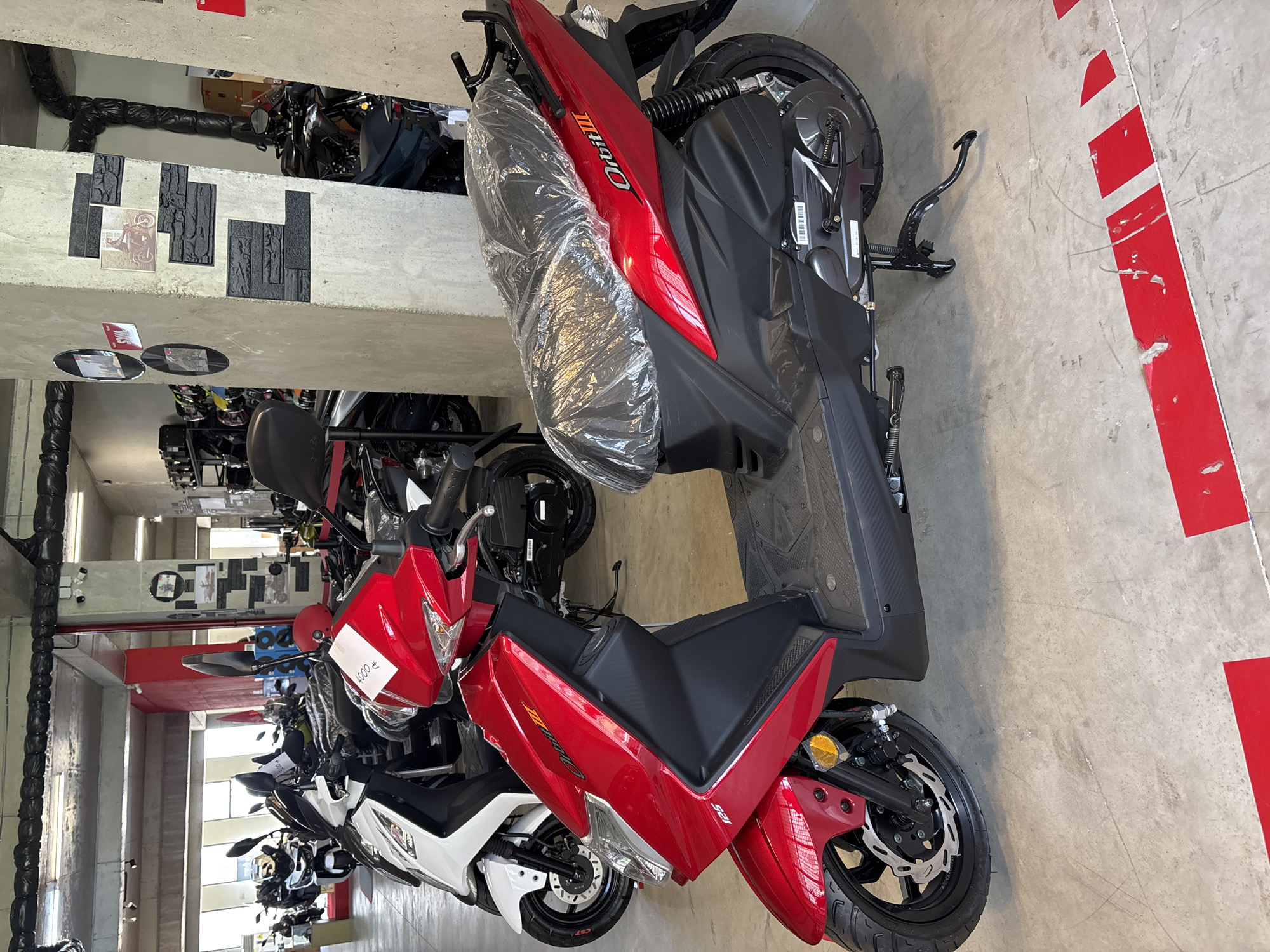 The image shows a red scooter parked indoors, likely in a showroom. The scooter is the primary subject, with its sleek design and reflective red bodywork prominent. It appears new, with plastic wrap covering the seat. The scooter has a black seat, handlebars, and wheels with disc brakes. The brand name "Orbit III" is visible on the side panel.
In the background, other scooters are visible, including a white one parked to the left, and darker models further back. The environment is an indoor space with a concrete floor marked with red and white lines. The walls are a mix of concrete and what appears to be decorative stone panels. Industrial lighting fixtures hang from the ceiling. There are also some shelving units with what seem to be motorcycle helmets.
The presence of multiple scooters and the showroom-like setup suggests this is a dealership or a similar retail environment. The lighting and the overall cleanliness of the space further support this. There are no people visible in the image. Based on the provided context, this scene is located in Tbilisi, Georgia.