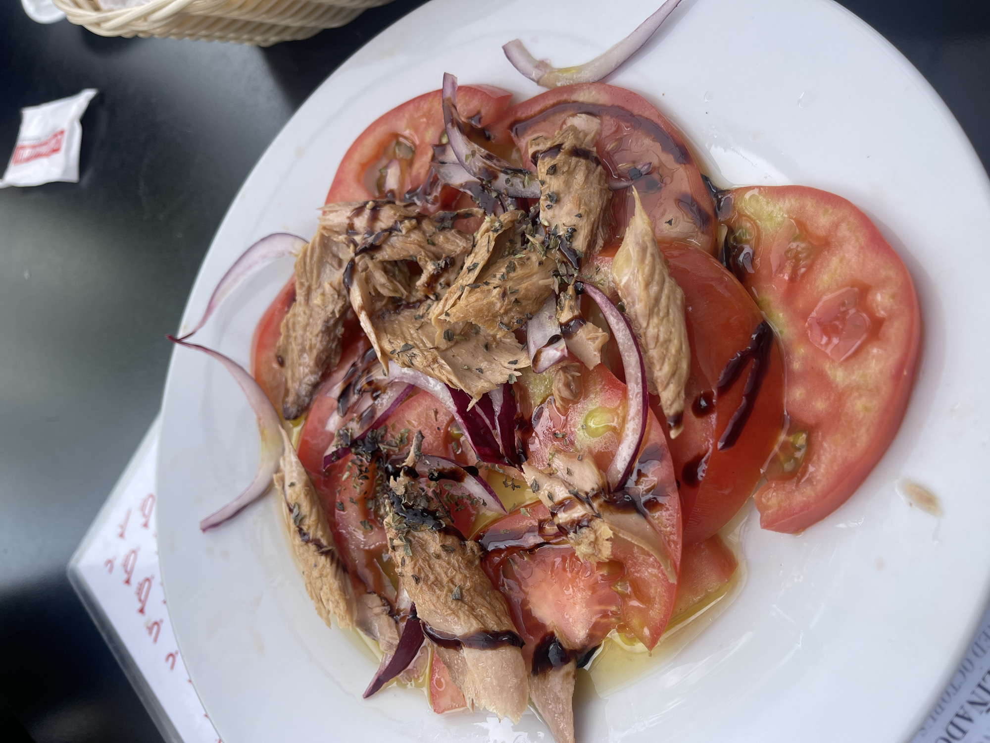 The image depicts a plate of food, specifically a salad with sliced tomatoes, red onion, and what appears to be canned tuna, drizzled with balsamic glaze. The plate is white and sits on a black table. A basket is visible in the upper left corner. A packet of sugar is also visible to the left of the plate. A menu can be seen on the lower right side. The food is centrally located in the frame and illuminated by ambient light. The setting appears to be an outdoor dining area in Jerez de la Frontera, Spain, based on the menu visible.