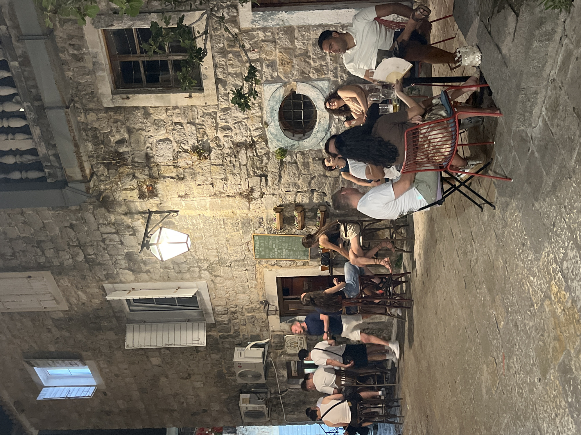 An outdoor dining scene is depicted at night in the old town of Budva, Montenegro. The image shows a stone building with several people seated at tables and standing at a bar. A woman in a white tank top and dark shorts is seated at a bar. To her left, a man in a white t-shirt and black shorts stands at the bar. Further left, two men in white t-shirts and dark shorts are standing. Next to them, a man in a navy t-shirt and white shorts is talking to someone. To the right, a woman with long dark hair is seated at a bar in a blue top and denim shorts. Further right, a group of people are seated at a table. A man in a white polo shirt and dark shorts is seated on the right side of the table. A woman with dark hair is seated to his left. Two other individuals are partially visible at the table. The environment is characterized by cobblestone pavement and a stone building facade. A lantern hangs on the wall, illuminating the area.