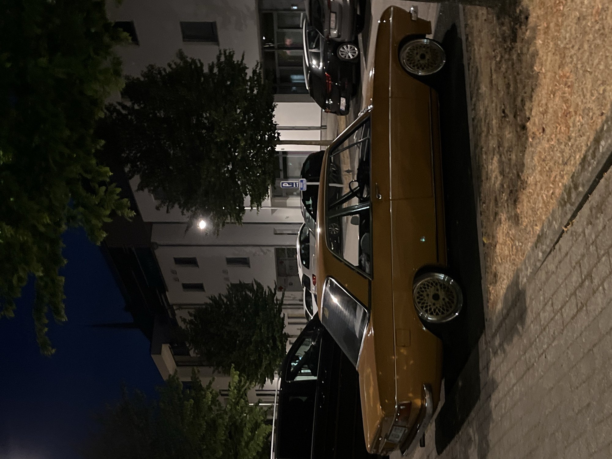 The image captures a nocturnal scene in what appears to be the city of Saint-Vith, Belgium, judging by the context provided. In the foreground, a vintage, golden-brown coupe is parked on a cobblestone surface. The car has distinctive chrome accents and custom mesh wheels. Behind it, several other vehicles are visible, including a black van, a white hatchback, and darker sedans or wagons. The background consists of modern, multi-story buildings with illuminated windows and large glass facades, suggesting a commercial or residential area. Trees with dense foliage line the street, their branches casting shadows. The overall atmosphere is quiet and urban, illuminated by artificial streetlights and building lights against a dark night sky.