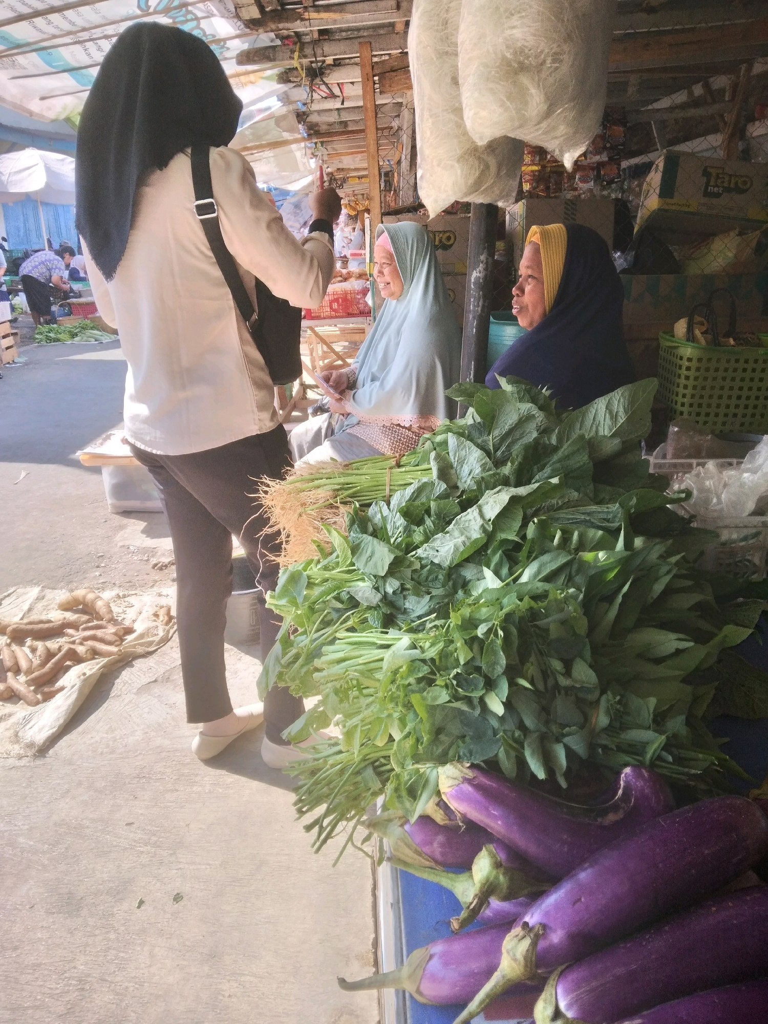 A medium shot captures an outdoor market scene in Sokaraja, Indonesia, bathed in sunlight. In the foreground, a generous pile of fresh, leafy green vegetables and several purple eggplants is displayed on a blue surface. To the left of the vegetables, a person wearing a black hijab and a cream-colored blouse stands with their back to the camera, facing two women. The woman on the left wears a pale green hijab and a patterned skirt. The woman on the right wears a dark blue hijab with a yellow trim. Both women are seated and appear to be engaged in conversation with the standing person. In the background, various market stalls are visible, with some produce like sweet potatoes laid out on a white tarp on the ground. Boxes and crates are stacked, and a green basket holds more produce. The overall impression is of a typical bustling market day.
