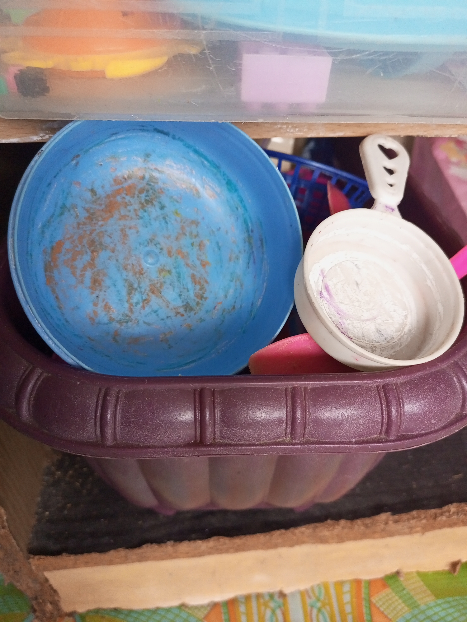 A close-up, high-angle shot shows a collection of plastic kitchenware items inside a cabinet. In the foreground, a ribbed, dark purple plastic bowl is visible, containing several items. A bright blue plastic basin with reddish-orange markings sits inside the purple bowl. To the right of the blue basin is a small, white plastic scoop with a heart-shaped hole in its handle, containing white powder with faint purple streaks. Behind these items, a portion of a blue plastic basket and a pink object are visible. The background reveals the interior of a cabinet with wooden shelves. The scene appears to be located in a domestic setting, likely a kitchen, given the presence of kitchenware. The lighting is dim, creating shadows that obscure some details of the environment. No people or specific actions are depicted.