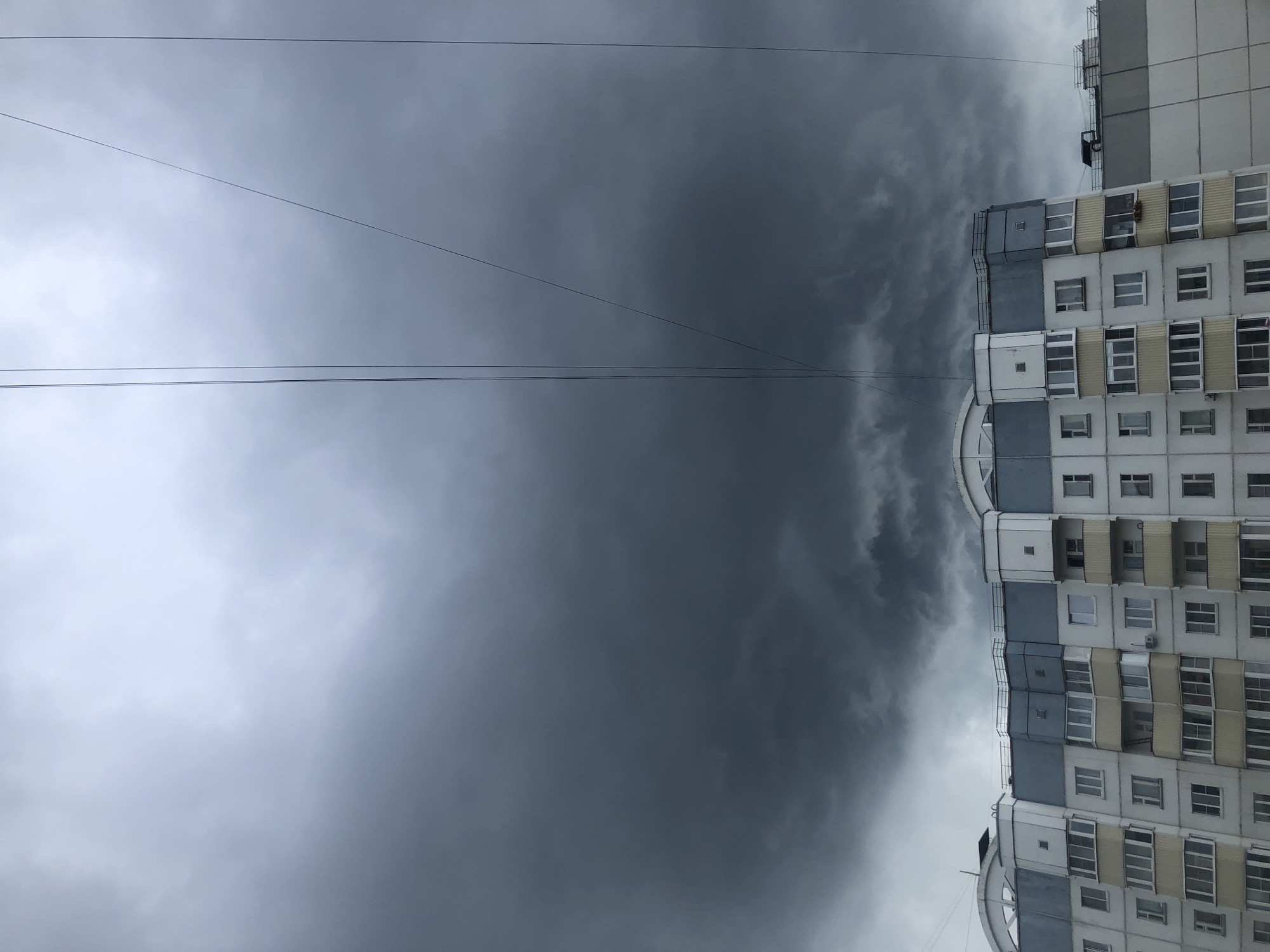The image shows a foreboding sky filled with dark, heavy storm clouds over a multi-story apartment building in Bor, Russia. The building is predominantly white with blue accents and yellow balconies. Several power lines are visible, stretching across the frame against the cloudy backdrop. The scene is captured from a low angle, looking upwards towards the sky and the upper portion of the building. There are no people visible in the image. The overall impression is one of impending bad weather.