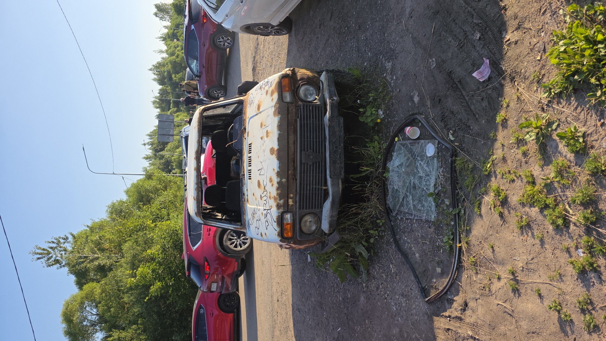 The media file shows a scene with several cars, some of which appear to be abandoned or damaged. The main focus is on a severely rusted white car in the foreground, with its hood open and some of its interior visible. A broken car window lies on the ground next to it. Several other cars, including red and white ones, are parked in the background.
The scene appears to be an outdoor setting, possibly a parking lot or a roadside area, with dirt and gravel on the ground, and some vegetation and trees. The time of day seems to be during daylight, possibly late afternoon given the lighting and shadows. The sky is clear and blue, suggesting fair weather.
There are some people visible in the background, but they are small and not the focus of the image. There is no discernible activity or event taking place; the scene appears static.
Notable details include the significant rust and damage to the white car, the broken window, and the presence of what looks like a crumpled piece of paper or fabric on the ground. Some graffiti is visible on the hood of the white car.
There is no readily discernible text in the image, other than what appears to be graffiti on the car. The overall impression is one of neglect and decay.