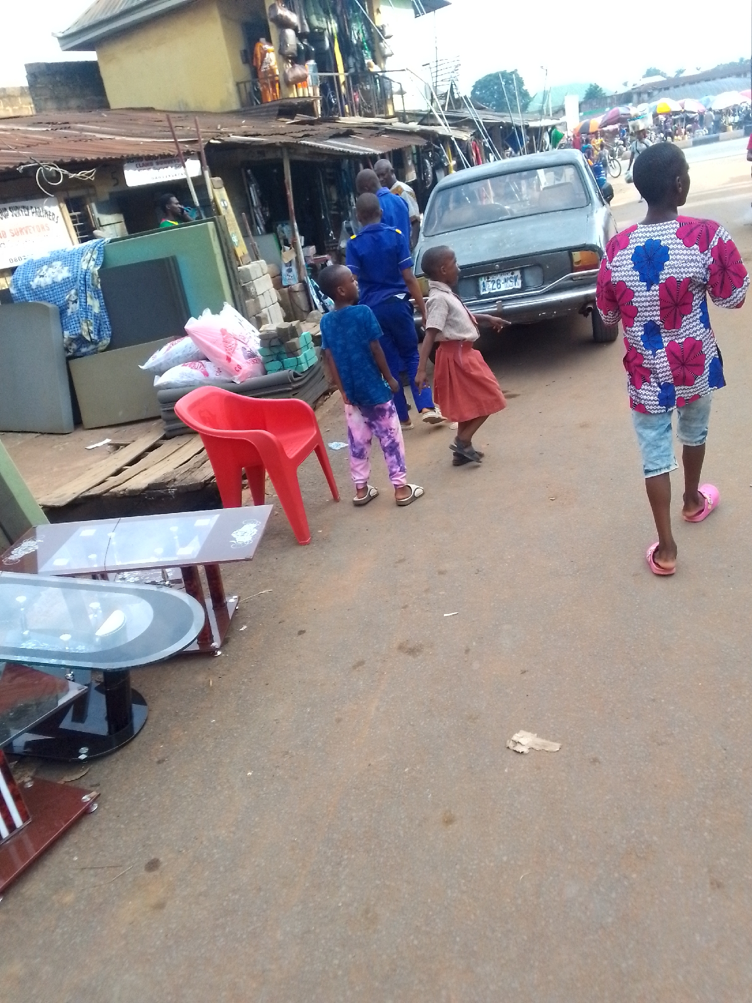 The image depicts a street scene in Orlu, Nigeria. There are several children present: one wearing a blue shirt and tie-dye pants, another in a school uniform of a beige shirt and reddish-orange skirt, and a third child wearing a pink and blue patterned shirt and jean shorts with pink sandals. There are also several adults wearing blue uniforms.
A light blue car with license plate A26HSY is in the background. There is furniture displayed, including tables with glass tops and a red plastic chair.
The setting appears to be a market area with buildings of varying construction and open storefronts with clothing or merchandise hanging outside. Other people can be seen in the distance. The street surface is a gray color.