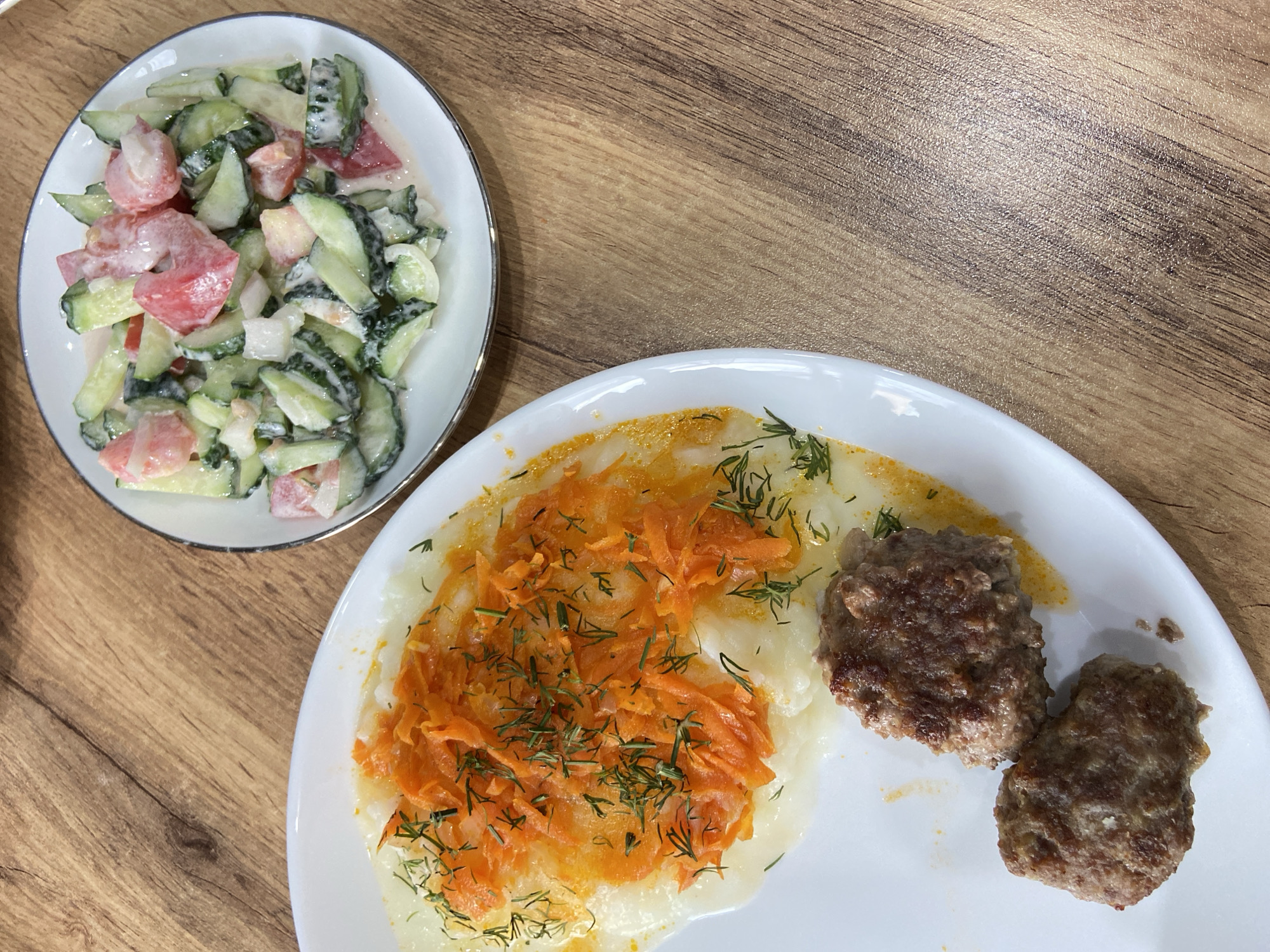 The image displays a close-up, top-down view of a meal served on a wooden table. On a large white plate in the foreground are two browned patties, likely meatballs or cutlets, resting on a bed of mashed potatoes. The mashed potatoes are topped with a generous portion of shredded carrots and garnished with finely chopped dill. To the upper right of the large plate is a smaller white plate containing a salad composed of sliced cucumbers, tomatoes, and possibly onions, all mixed with a creamy dressing. The wooden table has a visible grain pattern. The overall scene suggests a home-cooked meal. There is no specific indication of the city of Tujmazy, Russia, within the image itself.