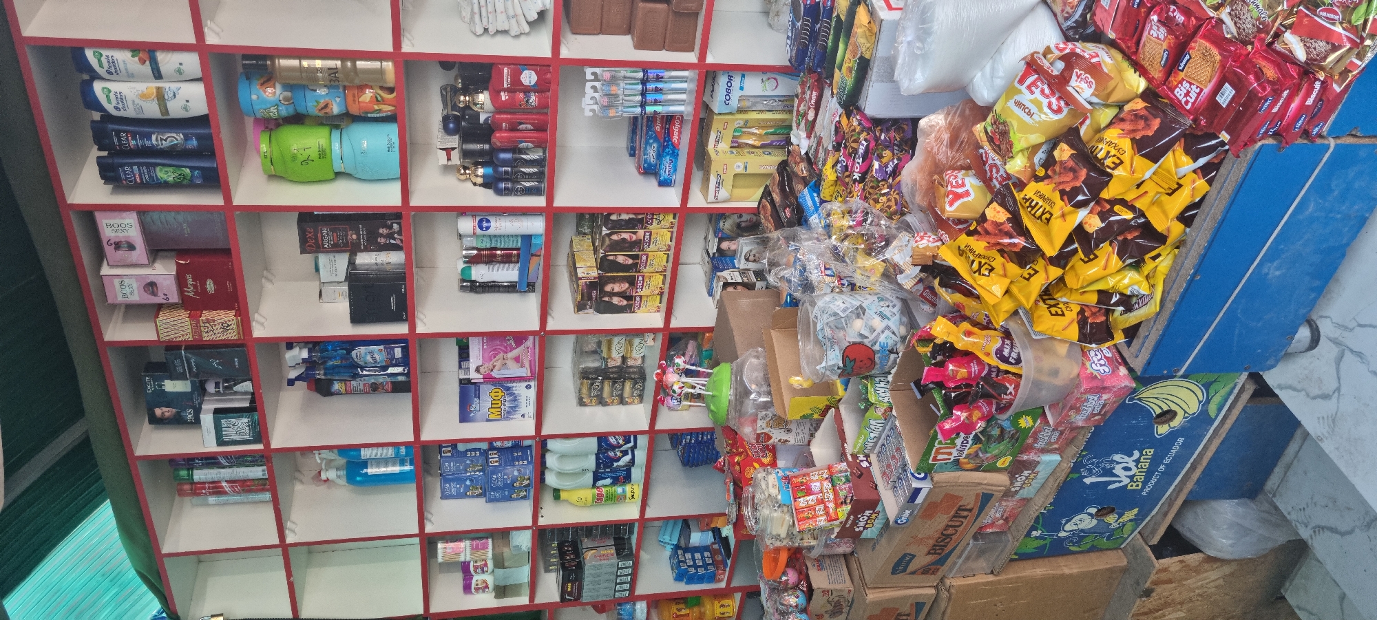 The image shows a shelf display in a shop, likely a small convenience store or kiosk, in Bokhtar, Tajikistan. The shelves are stocked with a variety of consumer goods, primarily hygiene products and snacks.
On the top shelves, various brands of shampoo bottles are visible, including Head & Shoulders and Clear. There are also boxes of cosmetics and perfumes, such as "BOOS SEXY" and "Marquis". Other items include spray cans, likely deodorants or air fresheners, and smaller packaged goods.
The middle shelves display a mix of items. One shelf has bottles of cleaning supplies or detergents. Another prominent section contains boxes of what appear to be hair dye or treatment products, with images of women on the packaging. There are also small boxes of biscuits and other snack items.
The lower shelves and counter area are overflowing with a large assortment of packaged snacks. Yellow and brown bags of chips or crackers are piled high, along with various smaller candy and biscuit packets. Jars filled with individual candies or small snacks are also present, along with some boxes of biscuits. A blue display box with the branding "Joe Banana PRODUCT OF ECUADOR" is visible on the bottom left, featuring illustrations of a monkey and bananas.
The environment appears to be indoors, with a green corrugated roof visible at the very top of the frame. The lighting suggests an artificial light source. The overall impression is of a busy retail space with a wide range of affordable goods. There are no people visible in the image.