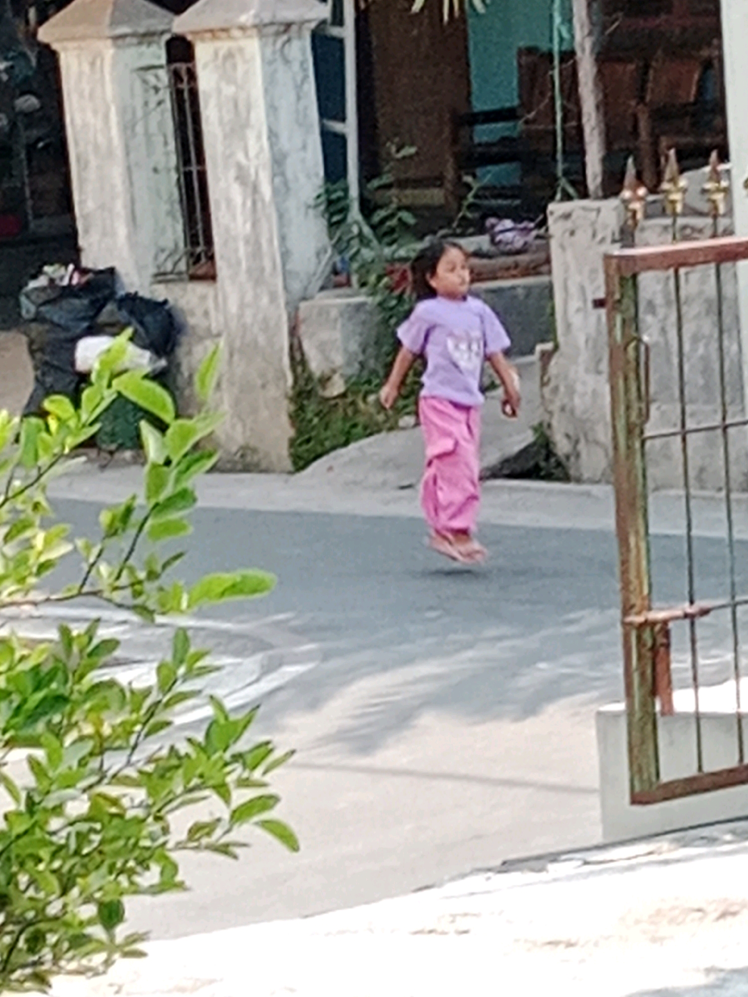 In Waru, Indonesia, a young girl, possibly of Southeast Asian descent, is captured mid-jump on a paved surface, likely a road or pathway. She is wearing a lavender t-shirt with a graphic design and loose-fitting pink pants. Her feet, clad in sandals, are elevated from the ground, suggesting she is engaged in an energetic activity, possibly jumping rope or dancing. She is facing towards the left of the frame with her gaze directed slightly downwards and to her left.
The background reveals elements of an urban or semi-urban environment. To the left, two decorative concrete pillars stand, with a dark-colored pile of trash bags situated at their base. Lush green foliage from a leafy plant drapes down in the foreground, partially obscuring the scene. Behind the girl, a portion of a building is visible, with a barred window on the left structure. A metal gate, with visible rust and vertical bars, occupies the right side of the image, with a concrete post supporting it. The ground surface is asphalt or a similar paved material, with subtle shadows suggesting overhead lighting or structures. The overall lighting indicates it is daytime.