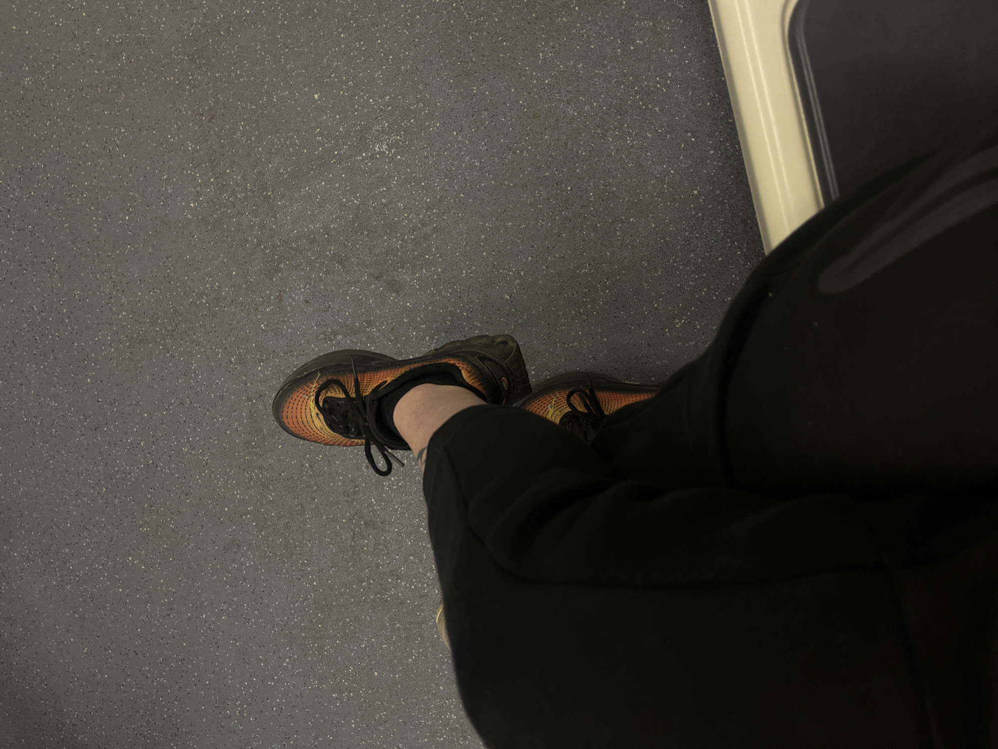 The image shows a first-person perspective of someone sitting on what appears to be public transportation, likely a train or bus. The person is wearing black sweatpants and orange and black running shoes. The floor is a speckled grey. Part of a white panel, possibly a window frame, is visible.