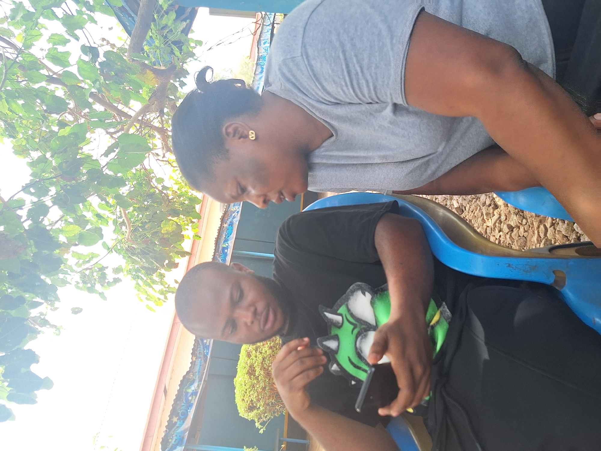 This outdoor photograph depicts two individuals, a man and a woman, in what appears to be a casual setting under bright daylight. The image is vertically oriented, showing the subjects from approximately the waist up.
On the left, a woman with dark skin and her hair pulled back is seen leaning forward. She wears a light grey short-sleeved top and what seem to be grey shorts or a skirt. A distinctive gold earring, shaped like a figure '8' or an infinity symbol, is visible in her right ear. She is looking downwards, her gaze directed towards the man.
On the right, a man with dark skin is seated on a blue plastic chair or bench. He wears a black short-sleeved t-shirt and black pants or shorts. He is holding a black smartphone in his left hand and a unique green, white, and black object, possibly a mask or a character toy, in his right hand. He is also looking downwards, focused on the items in his hands.
The background reveals a vibrant outdoor environment. Overarching tree branches with lush green leaves dominate the upper left portion of the image, allowing bright sunlight to filter through. Behind the seated man, a building structure with a light-colored roof and a blue-and-white patterned fascia is visible. Partially legible text, including "SHINE ON," can be seen on this trim. The ground beneath the seating area is covered with small, light-colored gravel or pebbles.
The activity suggests a quiet, focused interaction between the two, perhaps a shared moment of observation or a casual conversation. The time of day is clearly daytime, indicated by the strong sunlight. The overall atmosphere appears relaxed and informal.