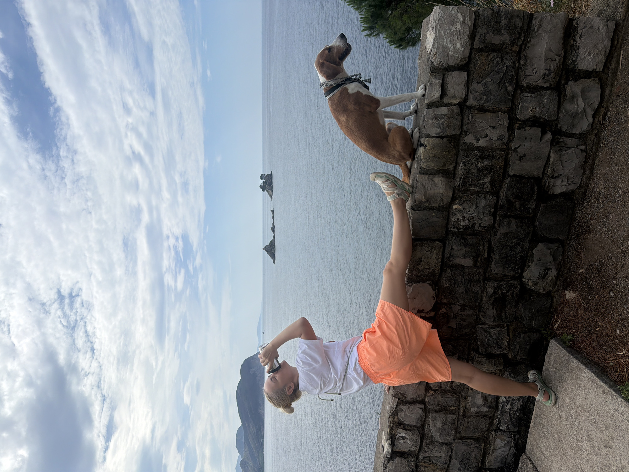 A woman and a dog are posing on a stone wall overlooking the Adriatic Sea near Golubovići, Montenegro. The dog, brown and white with a patterned collar, stands on the wall looking towards the sea. The woman is bent over with her right leg straight, with her foot on the wall next to the dog. She is holding a camera up to her face with both hands, presumably taking a photograph. She wears a white shirt and orange shorts. A stone wall is beneath them, and the sea stretches to the horizon, with some rocky islets in the distance. The sky above is blue with scattered clouds. In the background are more mountains.