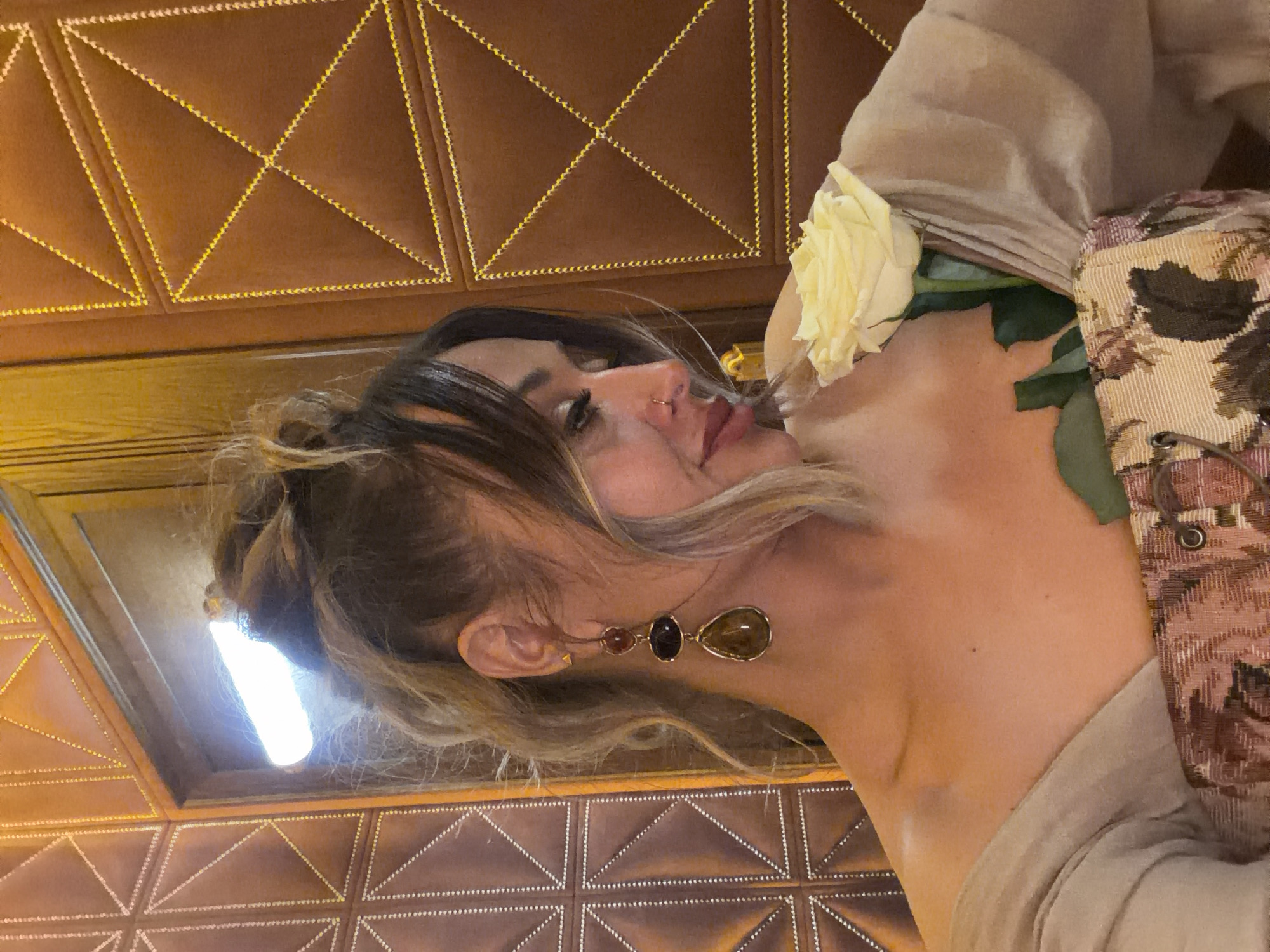 A woman with long brown and blonde hair is wearing large gem earrings and a beige top. A white rose with green leaves is positioned near her neck. She appears to be inside a building with a brown patterned ceiling. It is not possible to determine the exact location within Krasnoyarsk, Russia, based solely on this image.