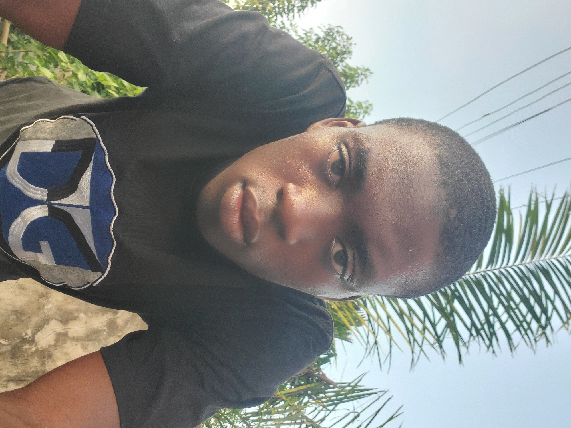 A young African man is taking a selfie outdoors, likely in Atani, Nigeria. He wears a black t-shirt with a blue and white design. The background includes a mix of greenery, palm fronds, and a pale sky. The perspective is from a low angle looking up at his face.