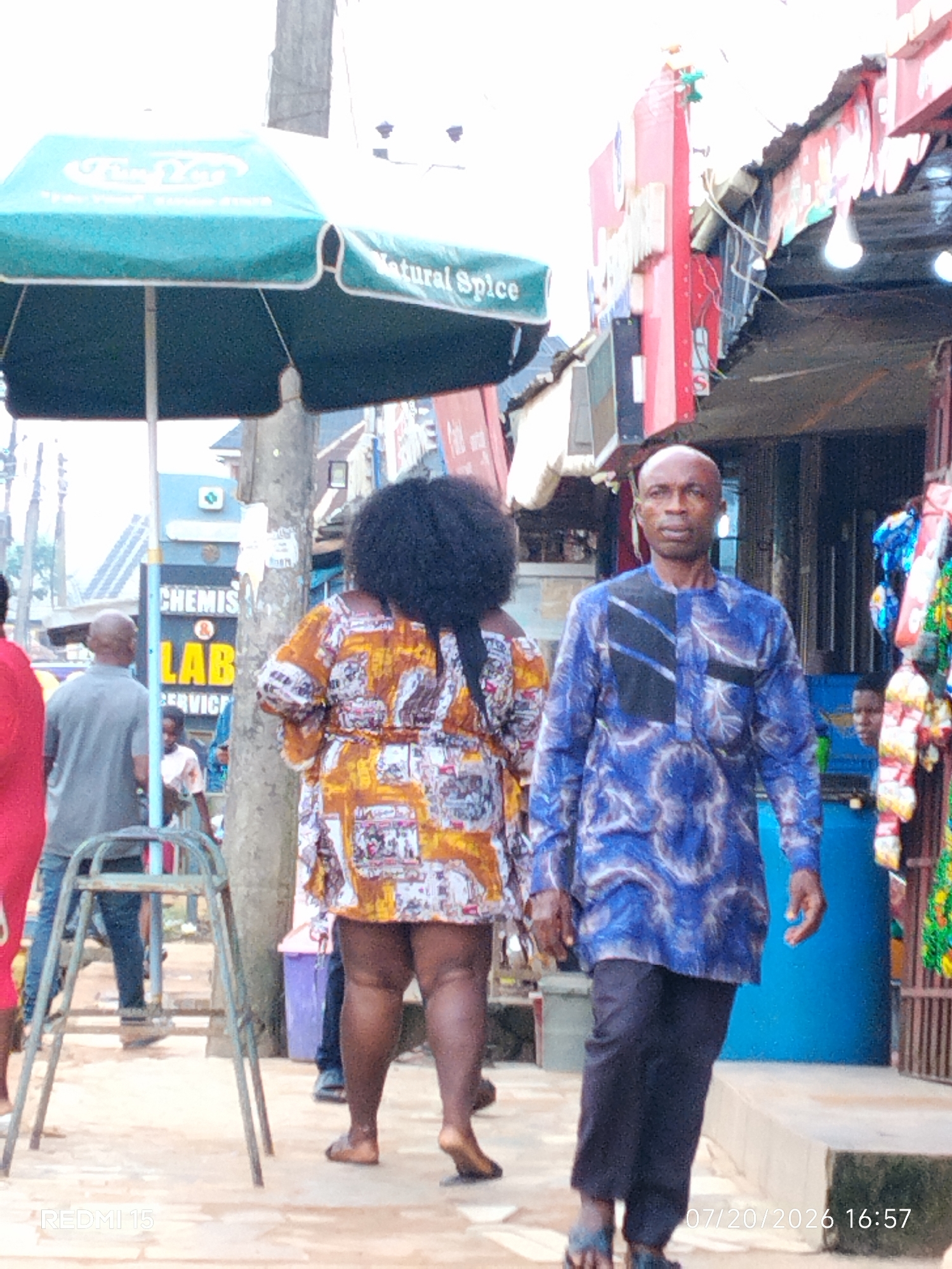In Umuode, Nigeria, a street scene unfolds under a teal awning with the text "Natural Spice" visible. A man in a blue and black patterned dashiki walks towards the viewer, his head bald and his expression neutral. To his left, a woman with voluminous dark hair wears a brightly colored, patterned dress. Behind her, partially obscured, is a sign for "CHEMIS & LAB SERVICE". Several other individuals are present in the background, including a person in a gray shirt and another wearing a red top. Various objects are scattered around the scene, such as a metal stool, a purple trash bin, and displays of colorful merchandise. The ground is paved. A timestamp in the lower right corner indicates "07/20/2026 16:57".
