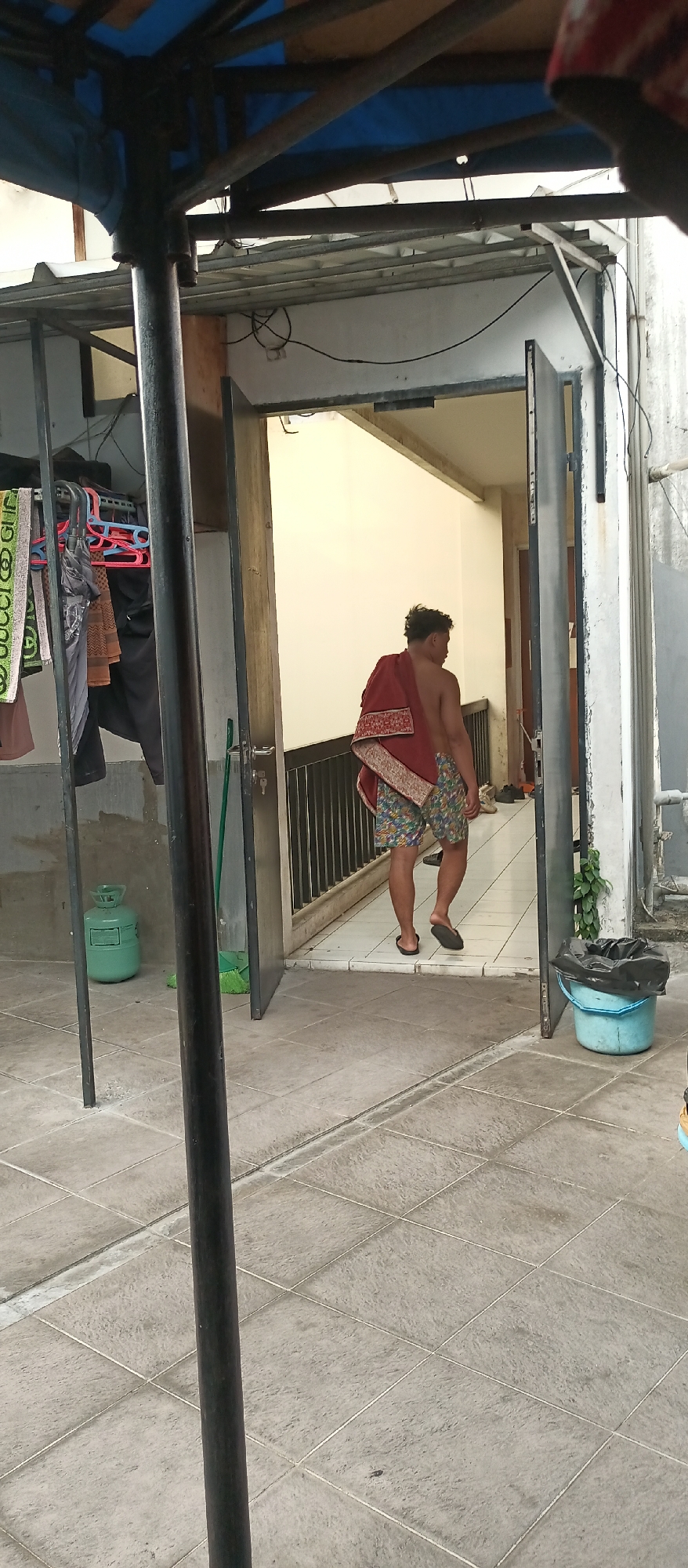 A person wearing patterned shorts and a maroon cloth draped over their shoulders walks through an open doorway on a tiled patio. The person appears to be moving away from the camera and towards the interior of a building. To the left of the frame, a green gas cylinder stands next to a clothes rack with various garments hanging on it. A dark-colored pole is prominent in the foreground, obscuring part of the scene. A blue bucket with a black trash bag is visible on the right side of the frame. The overall environment suggests an outdoor or semi-outdoor space in a residential or commercial area.