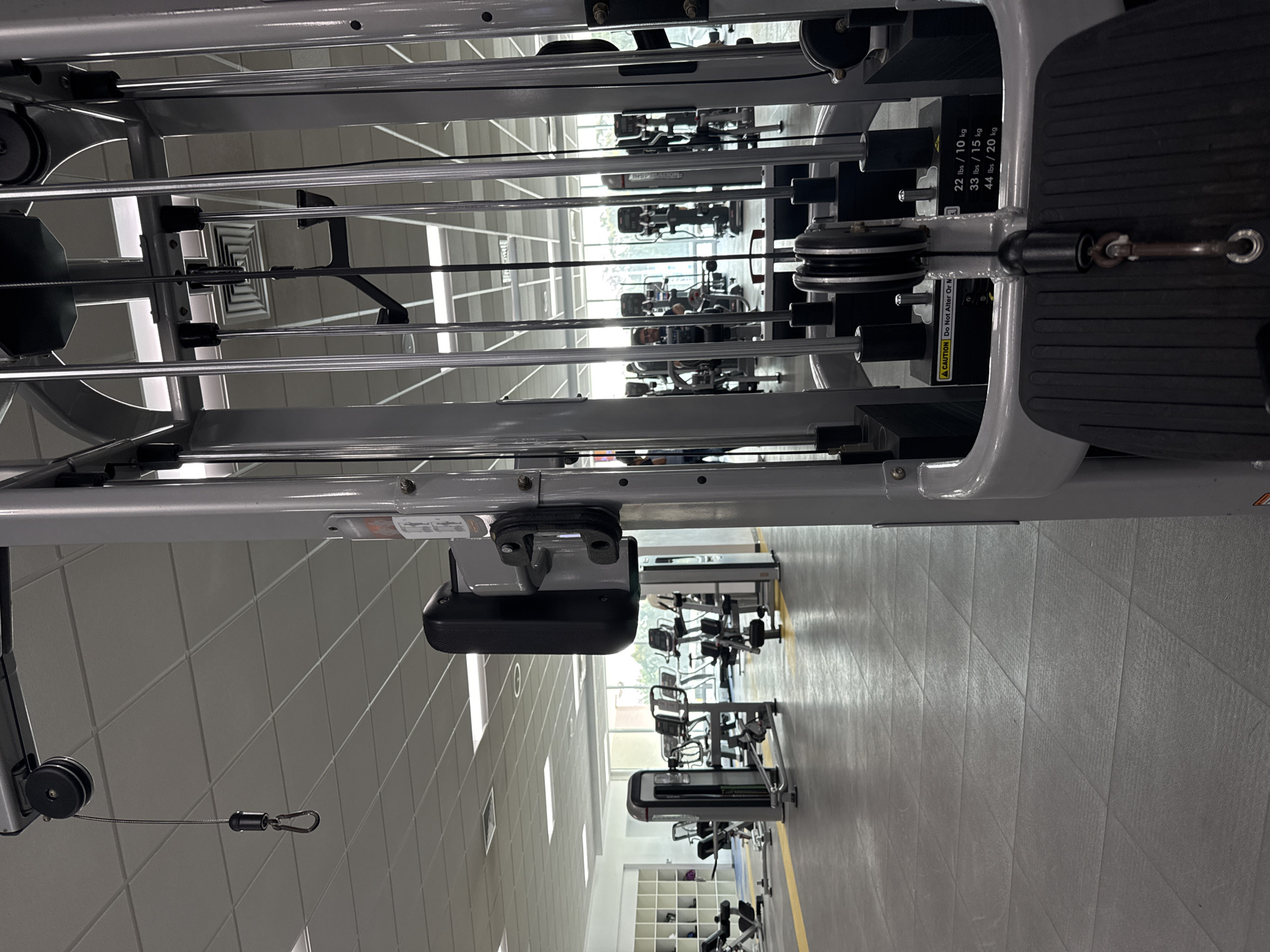 This image, taken from a low angle, showcases the interior of a gym, likely located in Heroica Puebla de Zaragoza, Mexico, given the image origin. The scene is dominated by rows of identical cable weight machines arranged in neat lines extending into the background. The machines' grey metal frames and black weight plates are visible, creating a repetitive pattern. The floor is covered in grey, geometrically patterned tiles, and a thin yellow stripe runs along the aisle. The gym's ceiling is tiled and illuminated by rows of fluorescent lights. Through a distant window, one can discern a glimpse of the outside environment. No people are explicitly visible in the immediate foreground, but faint figures can be seen in the background, suggesting that the gym is in use. The image composition primarily focuses on the equipment and interior architecture, capturing a sense of organized fitness space.