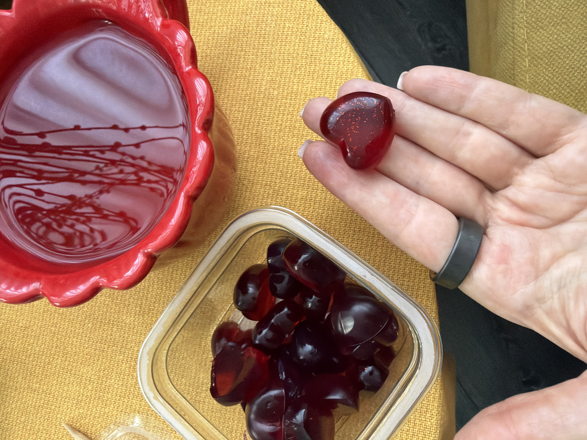 A close-up, eye-level shot shows a hand holding a red, heart-shaped gummy candy with a speckled texture. A container of similar gummy candies sits to the left, along with a decorative red bowl filled with liquid. A yellow textured surface serves as the background.
The scene does not appear to be in Moscow, Russia.