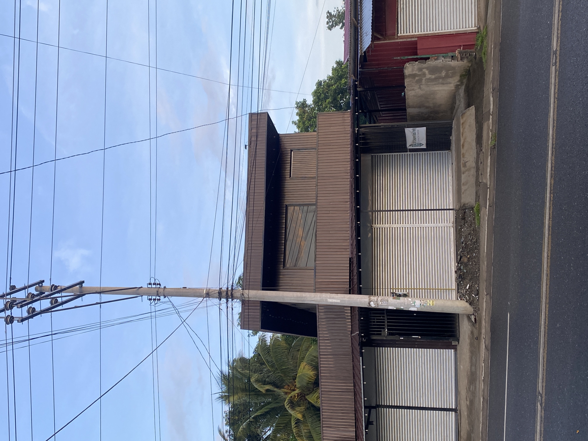 The image captures a street view in Davao City, Philippines, featuring a two-story building with a brown corrugated metal exterior. The lower level has several closed, silver roll-up doors, suggesting commercial establishments. A metal gate is visible between two of these doors. A sign with white lettering on a dark background is affixed to the building near the gate. The upper level of the building has a darker brown, angled facade with horizontal slats and a darker framed window. A utility pole with numerous power lines is prominently positioned in front of the building, extending into the sky. Palm trees and other lush green foliage are visible behind the building, indicating a tropical environment. The street in the foreground is paved with asphalt and has yellow lane markings. The sky is partly cloudy with a light blue hue. The scene appears to be a typical urban or suburban commercial area in Davao City.
