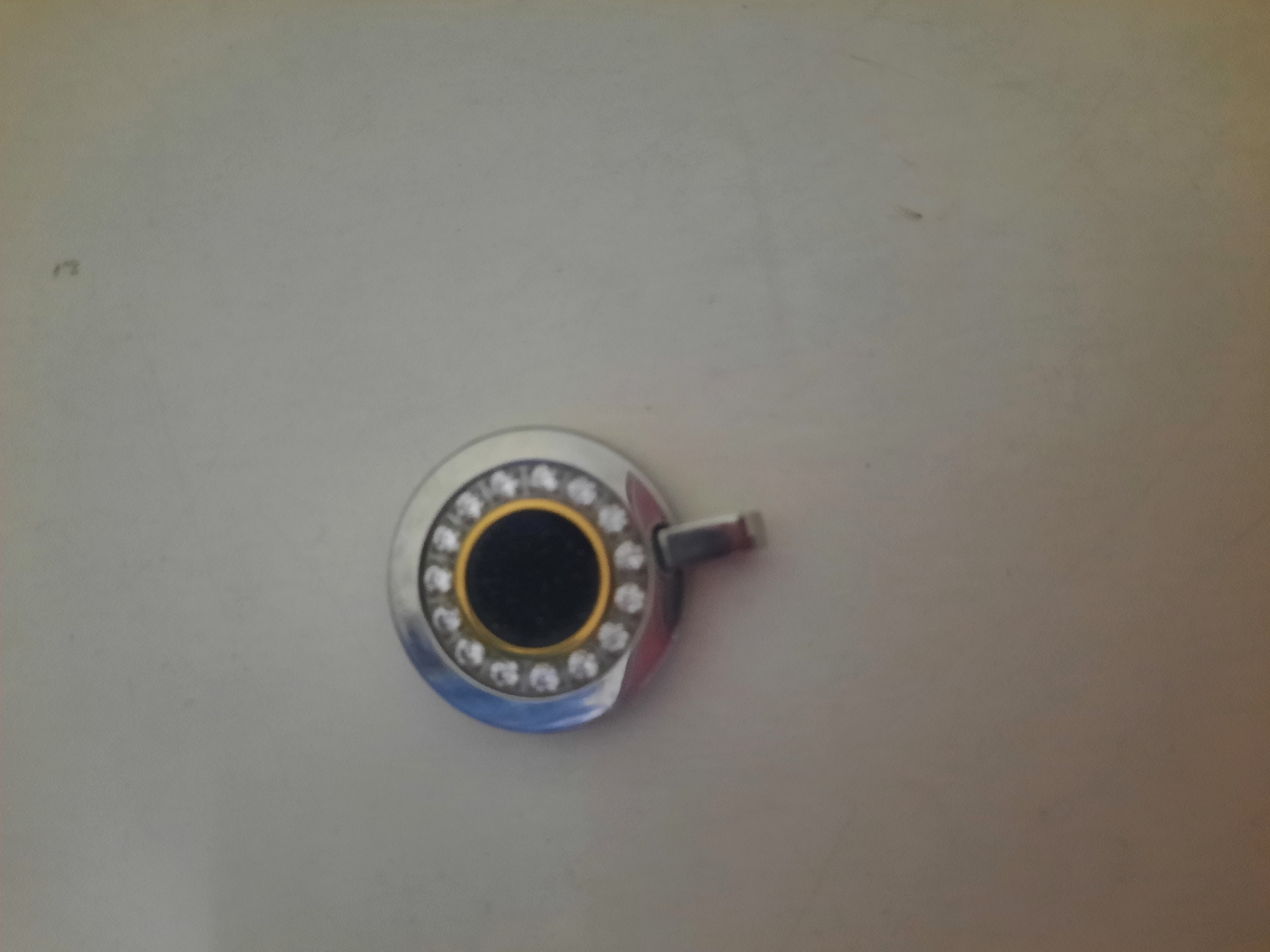 The image shows a round, silver-toned pendant with a black circular center. The outer edge of the pendant is adorned with small, clear stones, possibly diamonds or rhinestones, arranged in a circular pattern. There's a thin gold-colored rim around the black center. A small, metallic loop or clasp is attached to the bottom of the pendant. The pendant is lying on a light-colored, smooth surface, which appears to be a wall or a countertop. The background is mostly uniform and out of focus, with some subtle variations in lighting and tone. There is a faint, dark smudge or shadow on the surface to the left of the pendant. There are no people, animals, or other identifiable objects in the image. The lighting is soft and appears to come from above and slightly to the left, casting a very subtle shadow from the pendant. There is no indication of any specific location in Lhokseumawe, Indonesia.