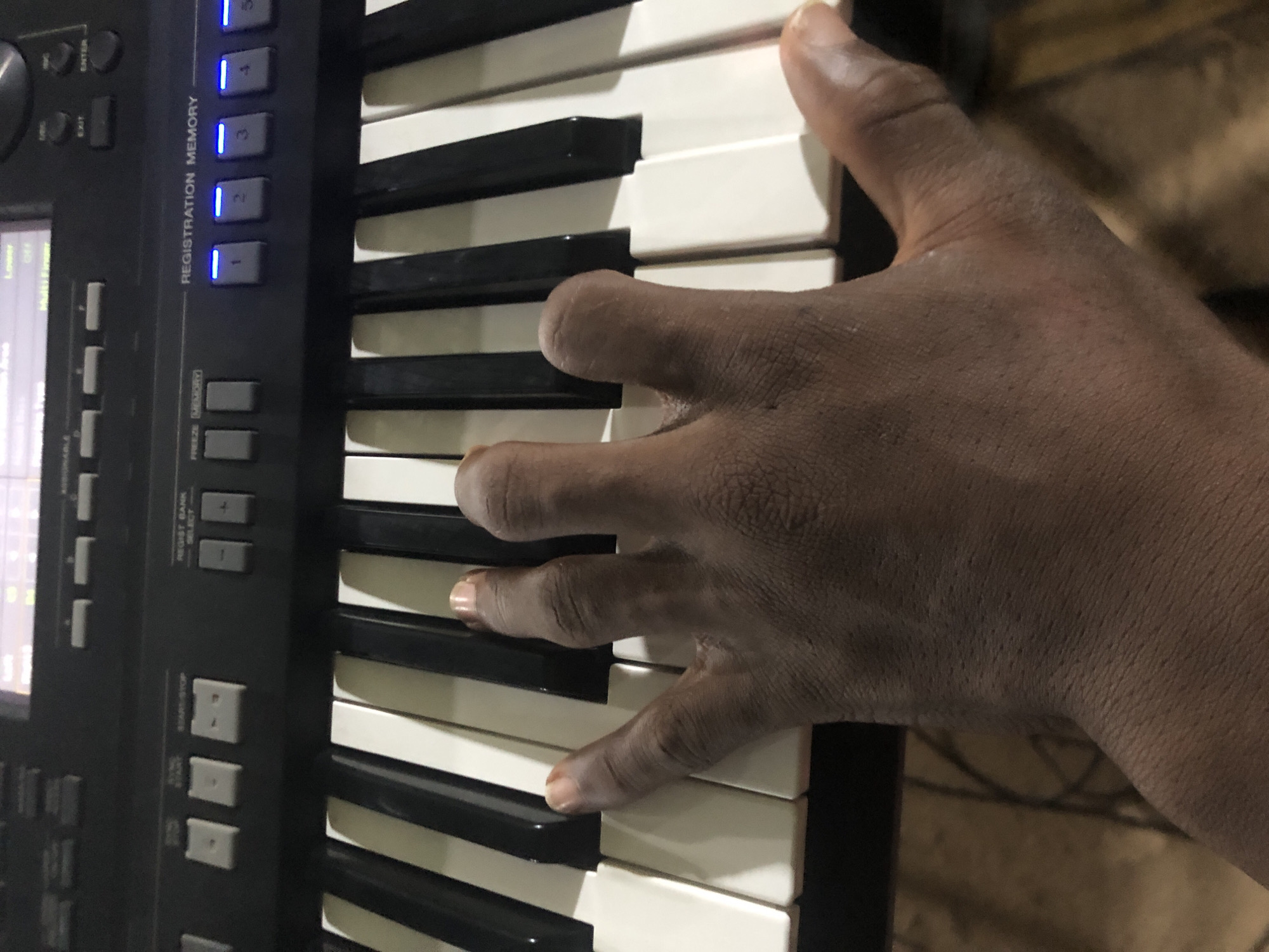A close-up image shows a hand playing a keyboard. The hand has dark skin and is positioned over the white and black keys. The keyboard has a control panel with buttons and a screen. The "REGISTRATION MEMORY" buttons are visible and illuminated with blue lights. The background is indistinct and appears to be a dimly lit interior. The location is not identifiable as Obosi, Nigeria.