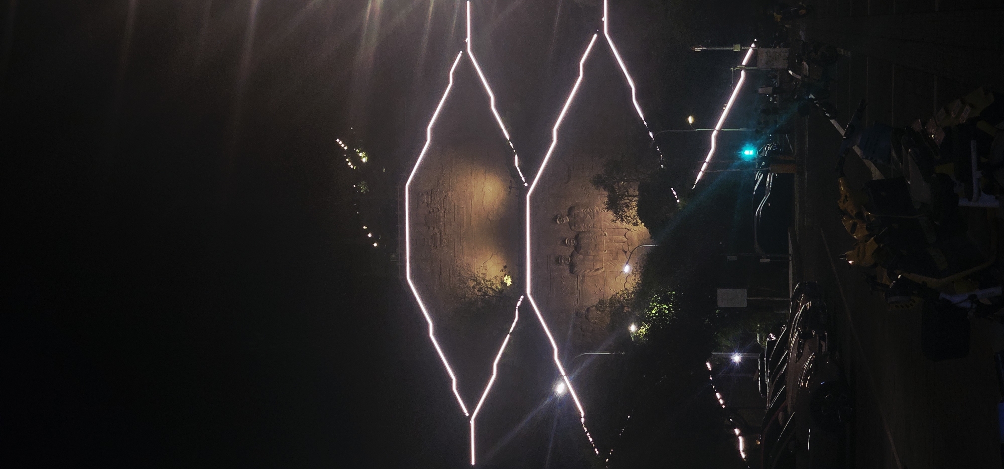 The image shows a nighttime view of a relief carved into a cliff face, illuminated by lights. The relief depicts three figures. The cliff is surrounded by trees and vegetation. White and pink neon lights are installed on the cliff face, forming geometric patterns. Below the cliff, a street is visible with parked cars and traffic lights. Several motorcycles are parked in the foreground. The scene is likely a public space in Beibei, China, possibly a tourist attraction or monument.