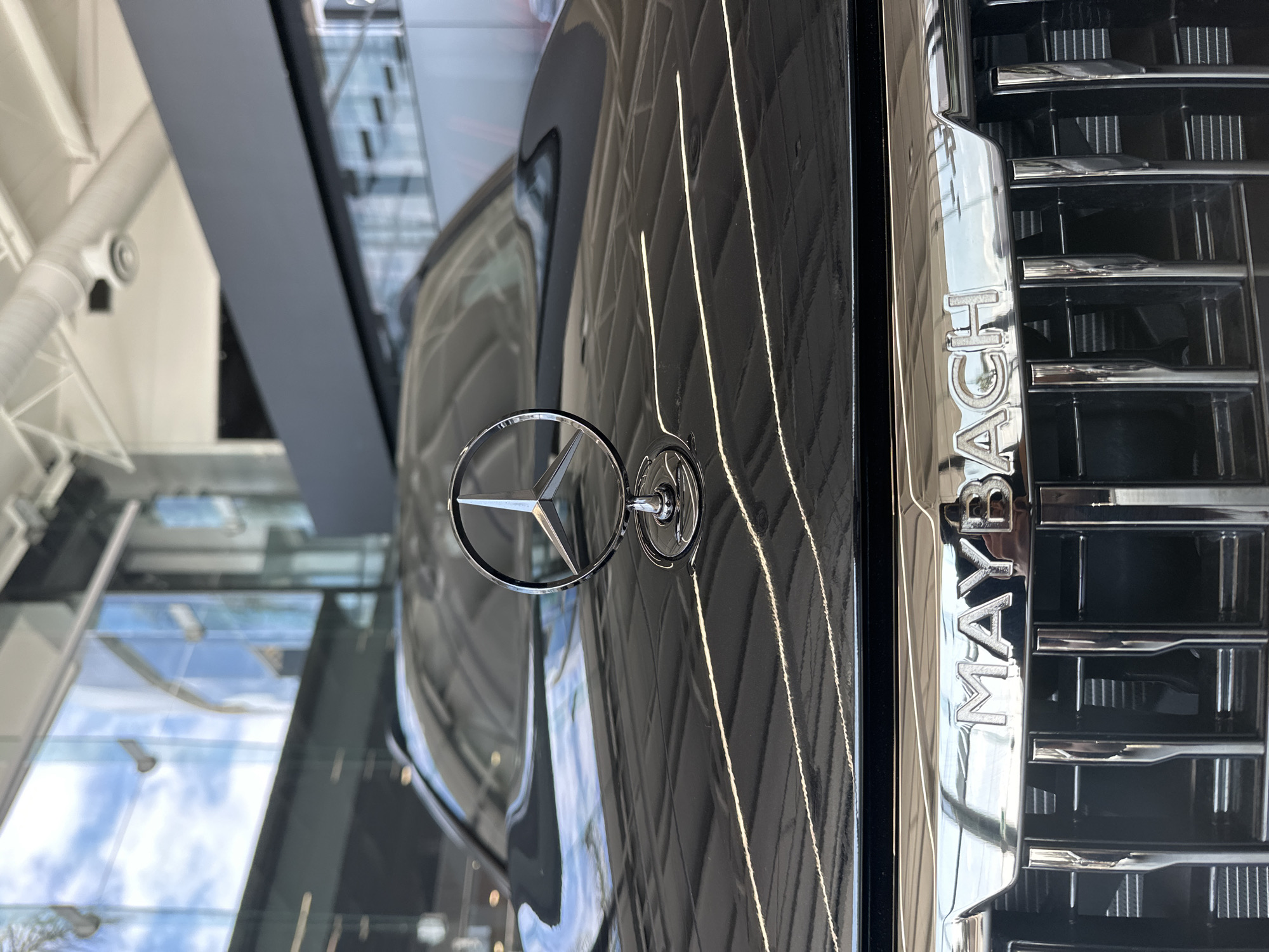 A close-up shot, taken at an angle, focuses on the hood and grill of a black Maybach car. The iconic Mercedes-Benz three-pointed star emblem is prominently displayed on the hood. To the right, the word "MAYBACH" is vertically spelled out on the grill. The reflections in the car's surface suggest the photo was taken indoors, possibly in a showroom or garage with glass walls, reflecting the sky and surrounding buildings. There is no information available from the image that locates it specifically in the city of San Bernardino Tlaxcalancingo, Mexico, or any indicators of people, actions, or interactions.