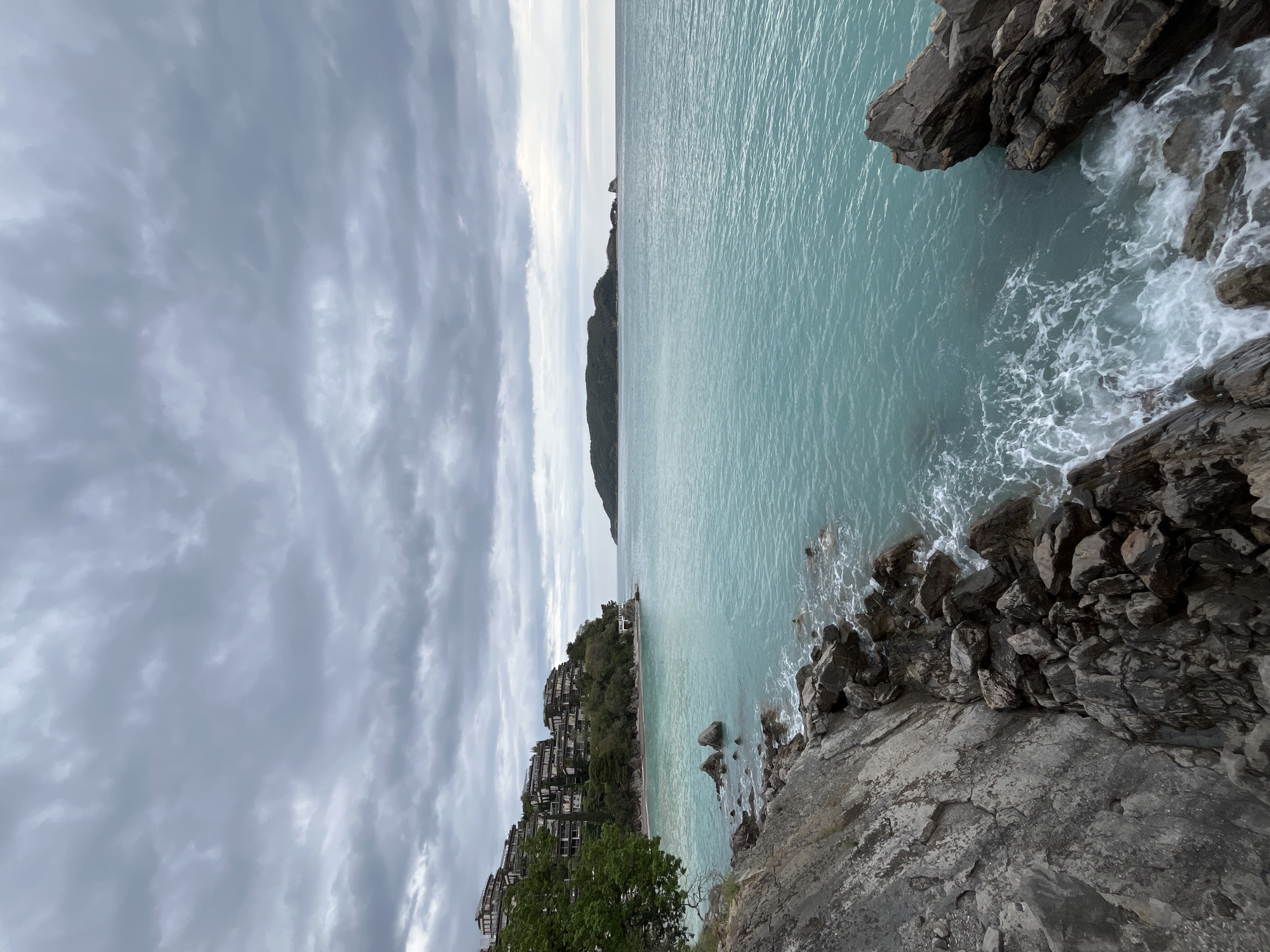 The image depicts a scenic view of the Adriatic coast near Budva, Montenegro, under an overcast sky. The azure sea is prominent, with white foam created by waves crashing against the rocky shoreline. The rocky terrain, composed of dark grey stones, juts out into the water. A stretch of coastline is visible, featuring buildings nestled among the vegetation on the hillside. In the distance, a promontory extends into the sea. The sky is filled with layered grey clouds, giving the scene a somewhat somber, moody atmosphere.