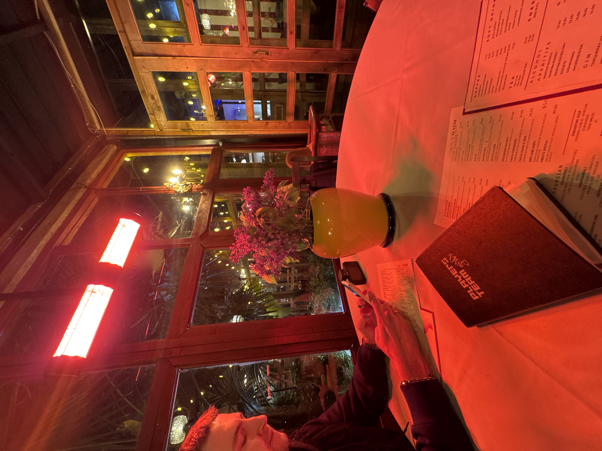 The image captures an indoor dining scene at a restaurant in Tbilisi, Georgia, likely in the evening or night. A person, visible in profile in the lower left, is seated at a table and appears to be looking at a menu. The table, covered with a white tablecloth, is brightly illuminated by a strong red light source, casting a warm, reddish glow across the entire foreground. On the table, there's a yellow vase holding a bouquet of purple flowers (lilacs).
Multiple menus are spread across the table. One menu, titled "MOVAMAISI", features sections like "STARTERS", "MAIN COURSE", "COFFEE", "TEA", "SOFT DRINK", "ALCOHOL", "APERITIFS", and notably, a "GEORGIAN" section with dishes like "CHASHUSHULI", "ADJAPSANDALI", and "CHIKHIRTMA SOUP". Another menu cover bears the text "PLAYERS TEAM girls".
The background features large wooden-framed windows, suggesting a conservatory-like setting or a room with expansive views. Through these windows, a dark outdoor environment with subtle, scattered yellow lights and foliage is visible. Two prominent, rectangular red lights hang in the upper left, contributing to the dominant red lighting scheme. The overall ambiance is intimate and moody due to the low light and strong color saturation.