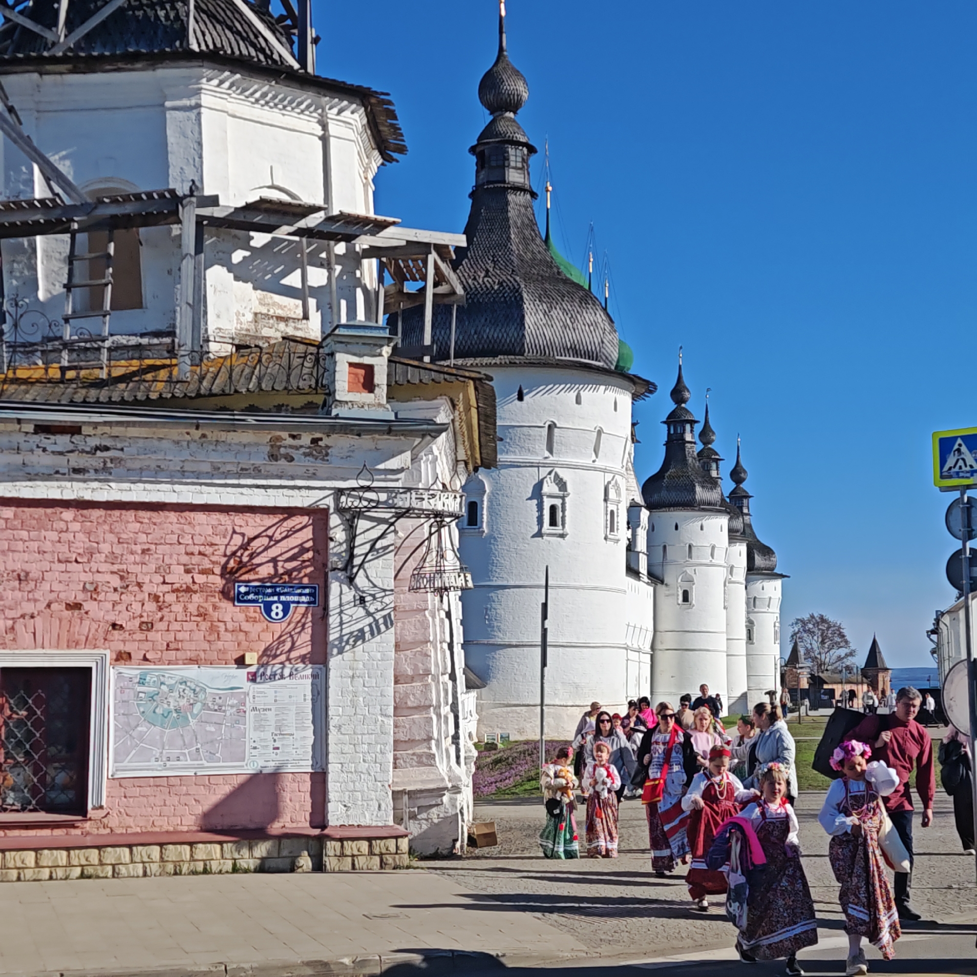 This vibrant image captures a sunny street scene in Rostov, Russia, offering a view of historic architecture and local activity. In the foreground on the left, a two-story building features a pink brick lower half with white trim and an upper section painted white, showing signs of wear and recent renovation with visible wooden scaffolding. A barred window is seen on its ground floor. On its pink wall, a blue street sign reads "улица Соборная площадь 8" (Cathedral Square Street 8), and an information board displays a map labeled "Ростов Великий" (Rostov Veliky, meaning Great Rostov) and "Музей" (Museum), confirming the location and hinting at the purpose of the grand structures nearby.
Dominating the right and background of the image are towering white stone walls and ornate, dark shingled onion-domed roofs, characteristic of traditional Russian ecclesiastical or fortress architecture, likely part of the Rostov Kremlin. Several of these domes are topped with spires, and some structures also show signs of scaffolding, suggesting ongoing preservation work. One dome is partially covered in green netting.
A group of people, including adults and several children, are walking on the paved sidewalk from right to left. Many of the children, predominantly girls, are dressed in colorful traditional Russian folk costumes, featuring patterned sarafans and decorative headwear, carrying items like small plush toys. This suggests participation in a cultural event, festival, or a visit to a historical site. Some adults accompany them, dressed in casual modern clothing, though one man wears a traditional-style red shirt. The sky is a clear, bright blue, indicating excellent weather, and strong sunlight casts distinct shadows, suggesting it is mid-day. A yellow and blue pedestrian crossing sign is visible on the far right.