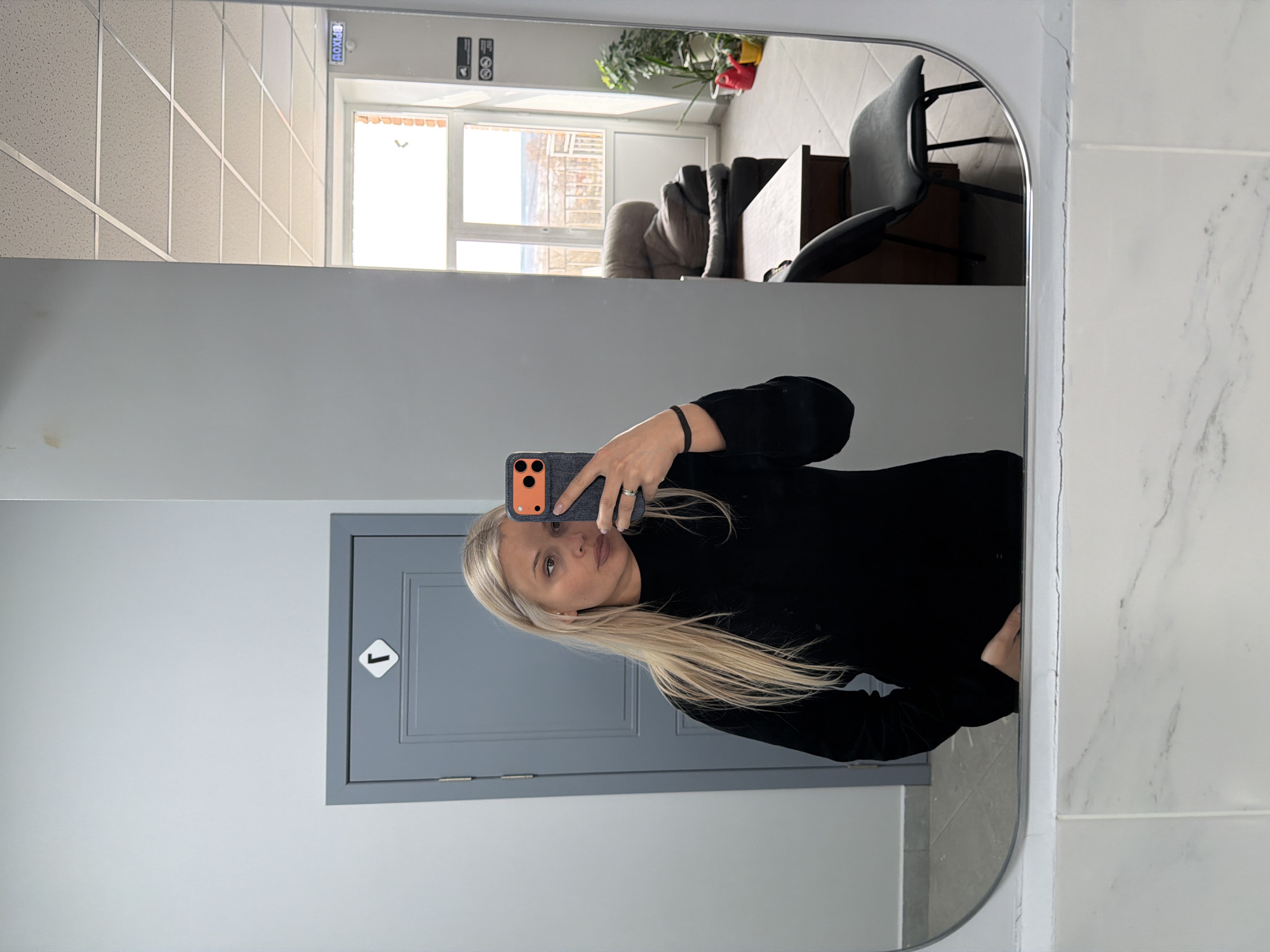 The image shows a woman taking a selfie in a mirror. The reflection reveals a room with neutral-toned walls and a gray door with a white symbol on it. Visible in the reflection is a portion of a room that includes furniture such as chairs and a couch. Outside the window, a glimpse of a cityscape can be seen.
While the location cannot be definitively confirmed as Sarapul, Russia, the prompt suggests this as a possibility. Without further evidence, the location remains unconfirmed.