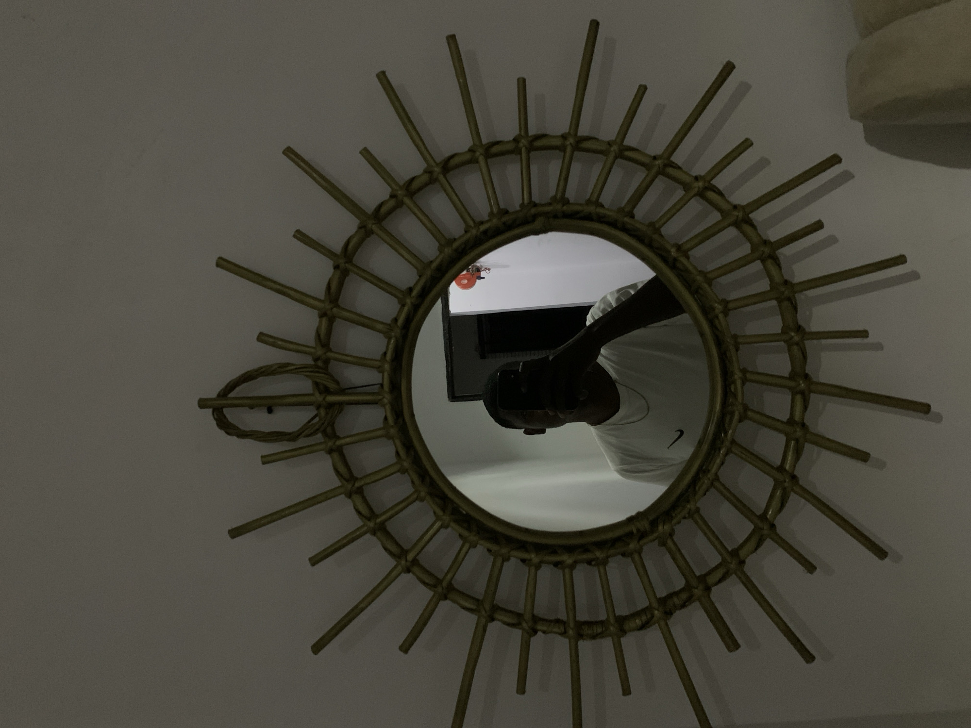 The photograph captures a gold-toned sunburst mirror mounted on a light-colored wall, likely within an indoor setting in Gwarinpa, Nigeria. The mirror's frame is intricately designed with numerous radiating "rays" made from what appears to be rattan or wicker, creating a decorative effect.
Within the circular reflection of the mirror, the upper body of the person taking the photograph is visible. The individual is wearing a white t-shirt featuring a prominent black Nike swoosh logo on the left side of the chest. Their head is also reflected, holding what seems to be a smartphone, presumably used to capture this image. Other reflections include a dark, rectangular object, possibly a television or monitor, and a small, circular orange object on the ceiling, which could be a light fixture or a smoke detector.
The scene is dimly lit, with soft shadows cast by the mirror's rays onto the wall, suggesting low light conditions, possibly evening or a room with minimal illumination. The primary activity depicted is the act of taking a self-portrait through the mirror. No explicit emotions are discernible.