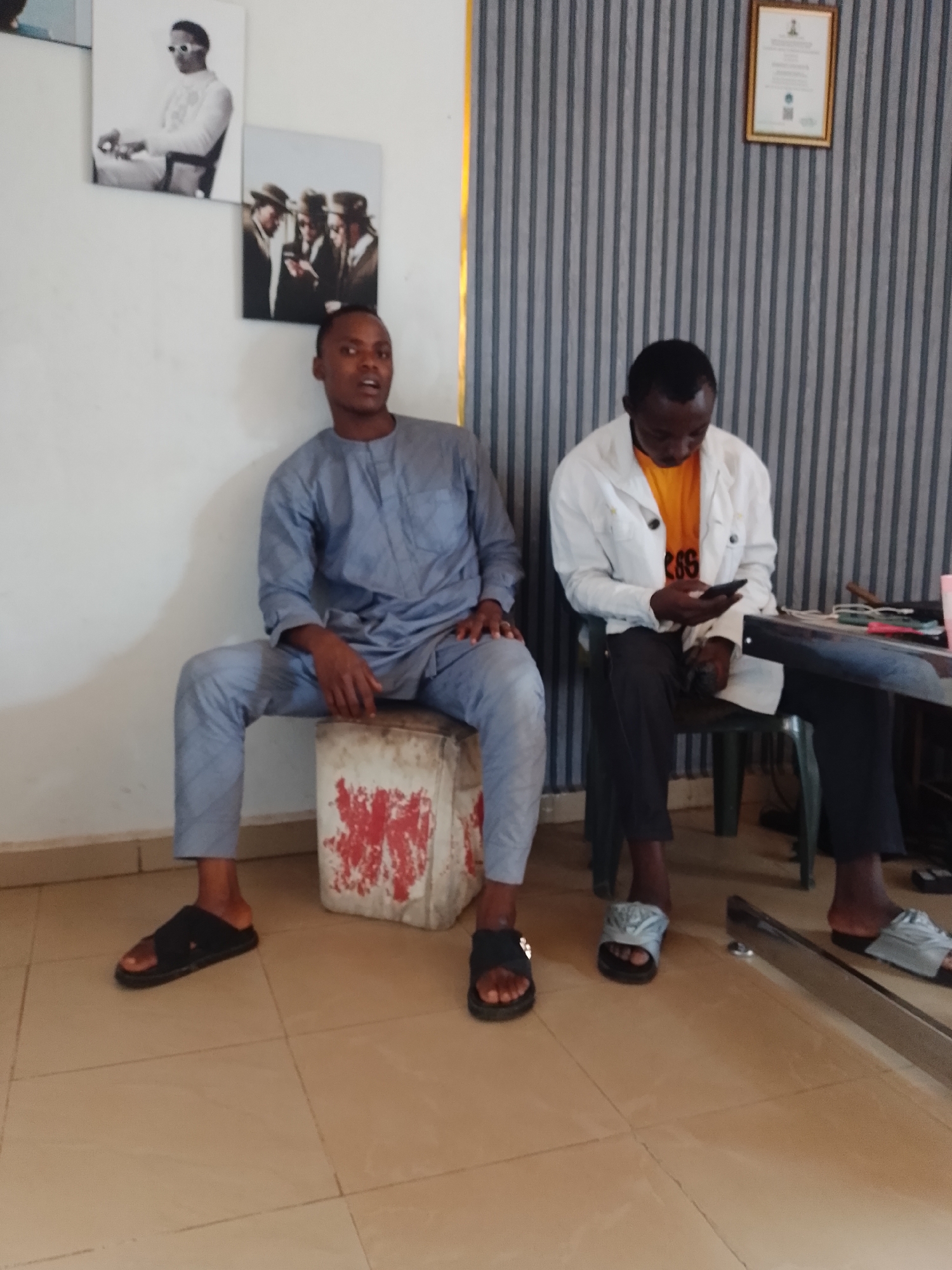 Two men are seated indoors, likely in a waiting area or office. The man on the left wears a light blue traditional outfit, consisting of a long-sleeved tunic and matching trousers, paired with black slide sandals. He sits upright on a white, red-stained block, looking directly ahead with a neutral expression.
The man on the right is dressed in a white jacket over an orange t-shirt, dark trousers, and light grey slide sandals. He is seated on a dark green plastic chair and is engrossed in his mobile phone, holding it with both hands and looking down. Partial numbers or letters, possibly "266", are visible on his orange t-shirt.
The background features a plain white wall on the left with two framed black and white pictures: an upper portrait of a man in sunglasses and a lower image of three men in hats and suits. On the right, the wall has vertical grey and white stripes, and a gold-framed certificate hangs high up. The floor is covered with light brown/beige tiles. A dark table is partially visible to the right of the man on the chair, with a pink cup and other small objects on it. The lighting suggests an indoor setting during the daytime.