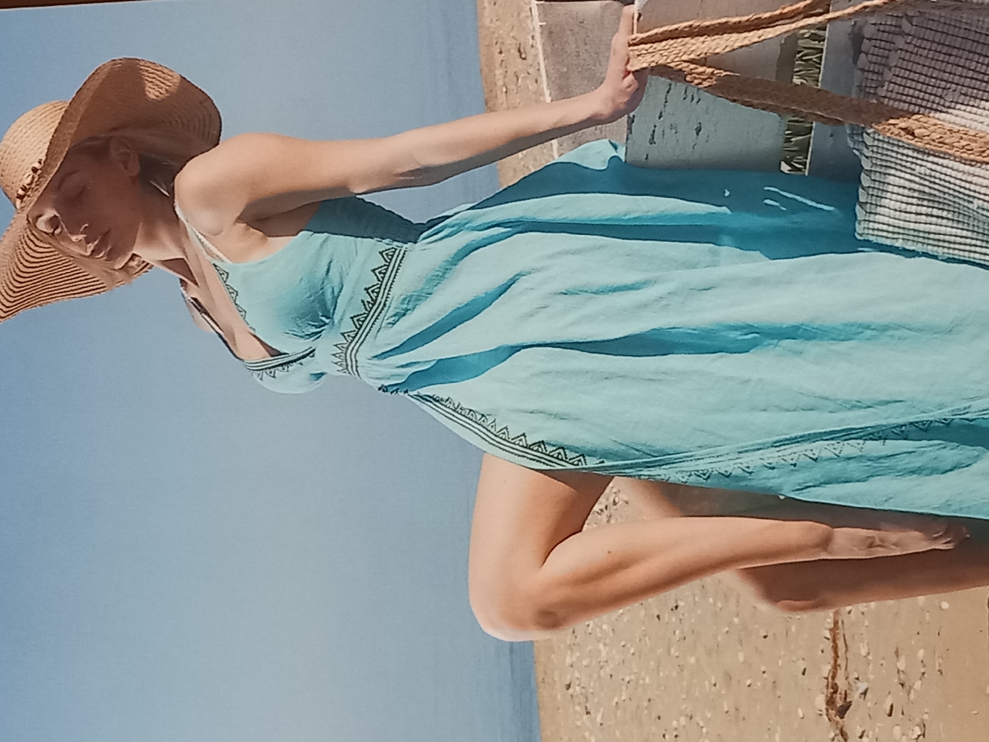 A woman with blonde hair, wearing a light blue dress and a straw hat, poses on a beach. She is seated on a low, white, weathered structure, with her left leg bent and her right leg extended. Her head is tilted back, and her eyes are closed. A woven straw bag with a striped interior is placed next to her right arm. The background consists of a sandy beach and a calm body of water under a clear blue sky.