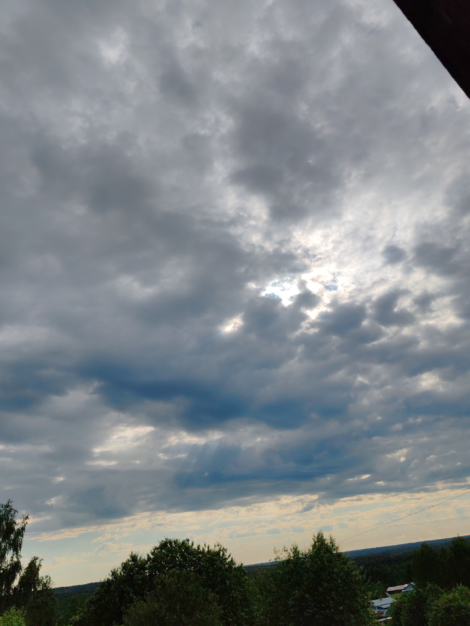 The image shows a cloudy sky over a wooded landscape. The sun is partially visible through the clouds, casting rays of light downwards. The sky is a mix of gray and blue, with dark storm clouds forming in the distance. The foreground consists of dense green trees, with some hints of houses visible in the distance. The scene appears to be a rural area, possibly on the outskirts of a city. The location is specified as Чухлэм, Russia, indicating a setting in the Russian countryside. There are no people or specific actions depicted in the image.