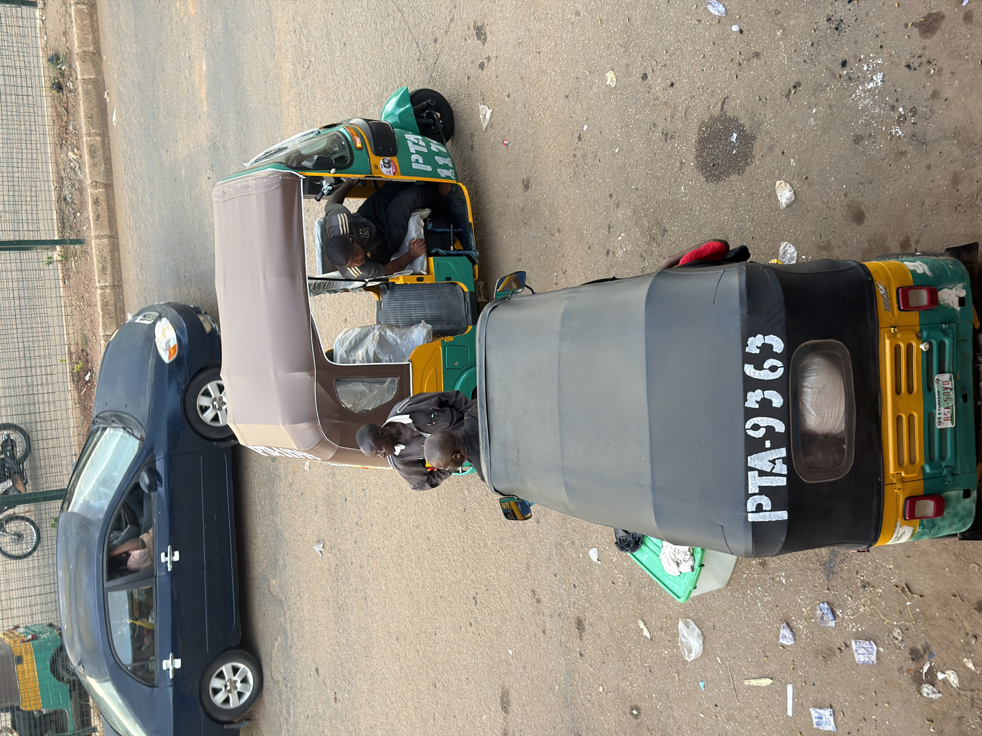 This image shows a street scene in Bukuru, Nigeria. The primary subjects are two auto-rickshaws, also known as Keke Napeps, and a dark blue sedan. One auto-rickshaw is in the foreground, its rear end prominent, displaying the license plate "PTA-936". It is yellow and green. The other auto-rickshaw is to the left, also green and yellow, with a person driving. This auto-rickshaw has the text "PTA 117" on its side. The blue sedan is parked on the left side of the frame, facing away from the viewer. A person is visible inside the sedan. The scene appears to be outdoors, on a paved road. There are some scattered pieces of trash on the ground. The lighting suggests it might be daytime, possibly late morning or afternoon, with no strong shadows indicating harsh sunlight. The overall atmosphere is that of a typical street scene in a Nigerian city, with vehicles and people going about their daily activities.