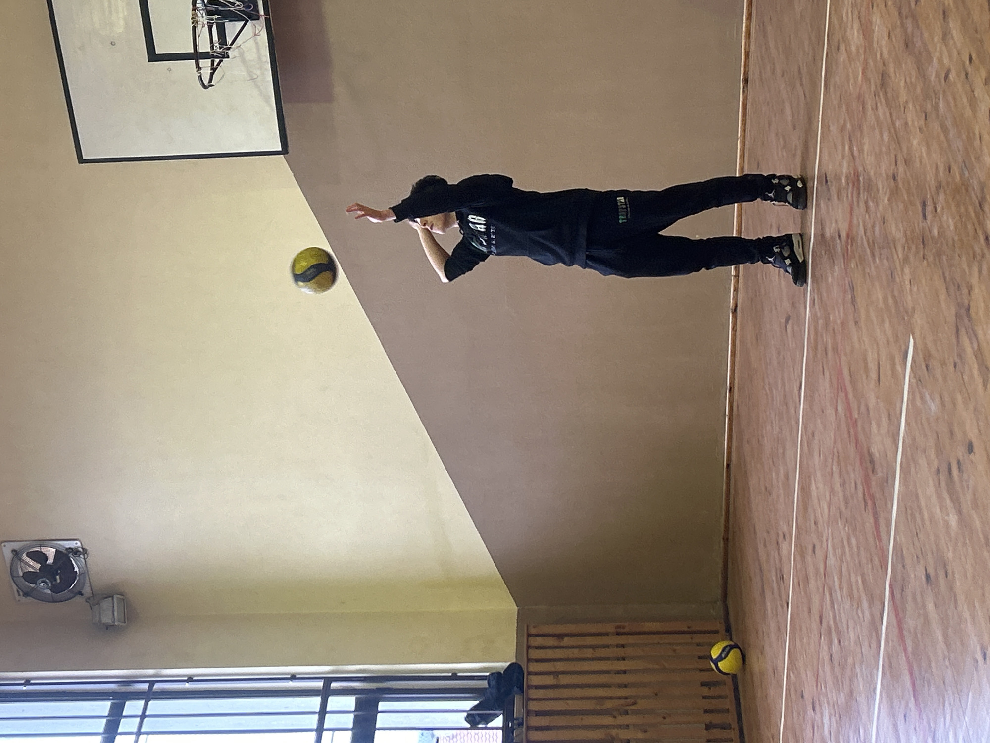 A young person, clad in black athletic attire, intercepts a yellow and black ball in a gymnasium in Zlatograd, Bulgaria. The gymnasium features a basketball hoop, a fan near a window, and wood floors with painted lane lines. Another similar ball rests on the floor near a wooden structure.