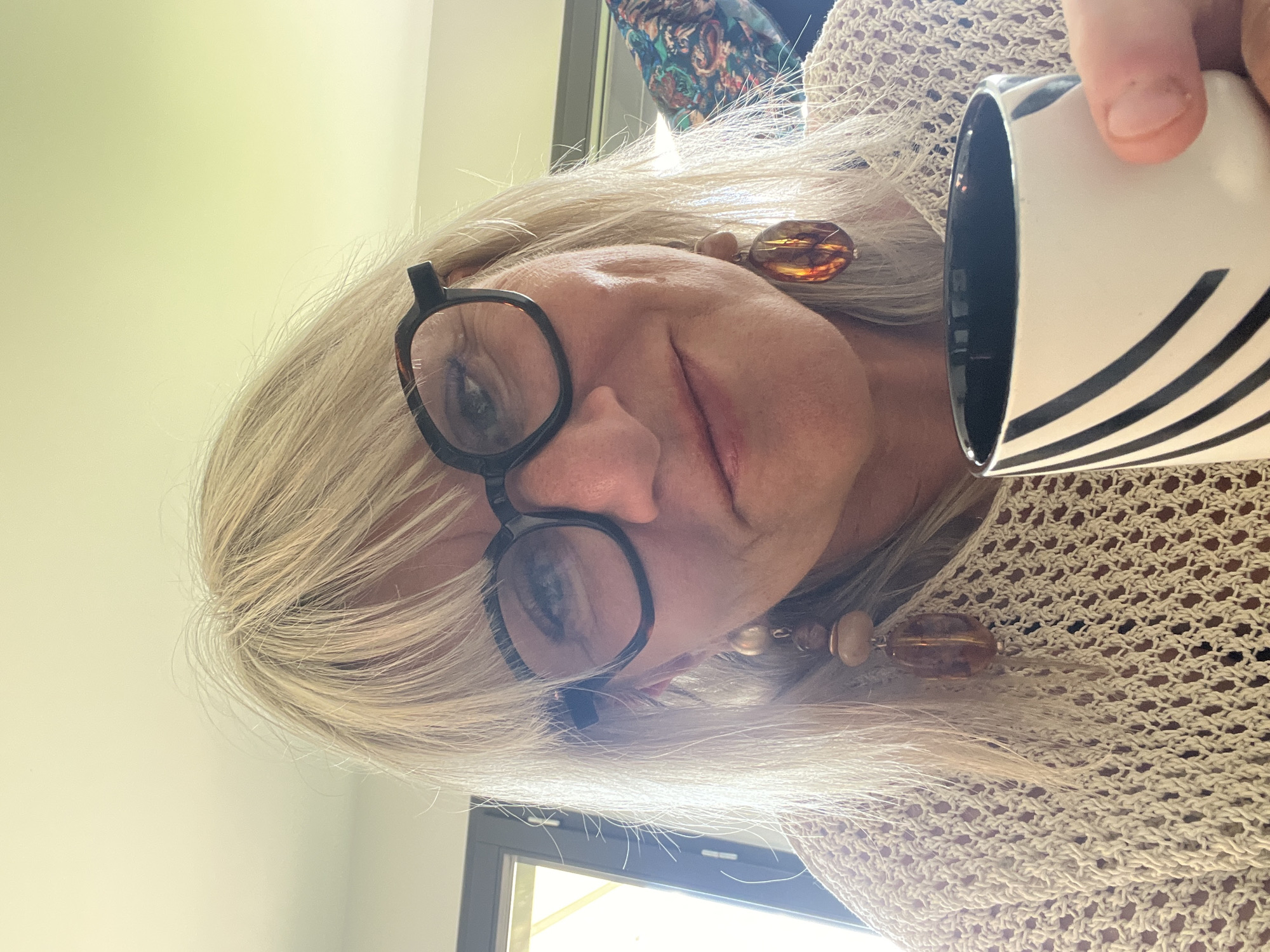 A person with blond hair and glasses is holding a black and white striped mug. They are wearing a beige crocheted top and amber-colored earrings. The environment appears to be indoors with a glimpse of a window and a pale green wall. There is no verifiable information about the location being Chambœuf, France, in the image.