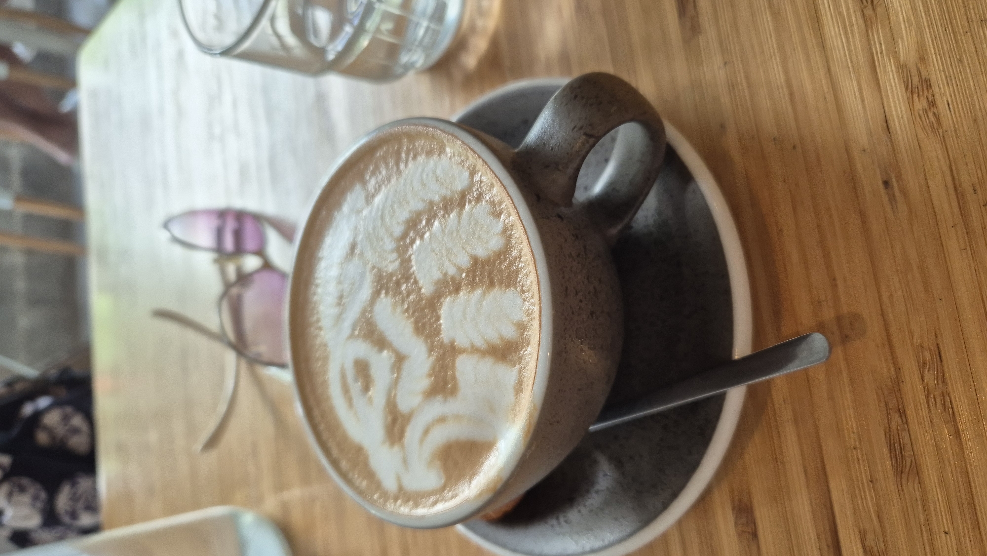 A close-up, high-angle shot shows a cappuccino with latte art on a wooden table. The latte art depicts a stylized animal, possibly a lion or a cat, with intricate swirling patterns. The cup, made of speckled gray ceramic, sits on a dark gray saucer. A metal spoon rests partially submerged in the coffee, angled across the saucer.
In the background, a pair of pink-tinted sunglasses with gold frames are casually placed on the table, slightly out of focus. To the right of the cup, a clear glass filled with water and ice is visible, also slightly blurred. Further back, a portion of a person's dark, patterned clothing is visible, suggesting they are seated at the table. The table itself is made of light-colored wood planks with a visible grain. The scene appears to be set outdoors, perhaps at a cafe, with the background suggesting an urban environment. The lighting suggests it is daytime.
This scene is likely located in Đà Nẵng, Vietnam, given the presence of outdoor cafes and the general ambiance often found in Vietnamese coastal cities.