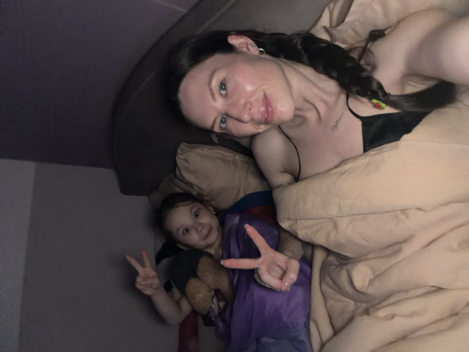 A woman with dark hair in a braid poses with a young child in a bed. The woman, wearing a black top, is covered in a beige blanket. The child, holding a teddy bear, is also in the bed with a purple blanket, making a peace sign with both hands. The room appears dimly lit and informal.