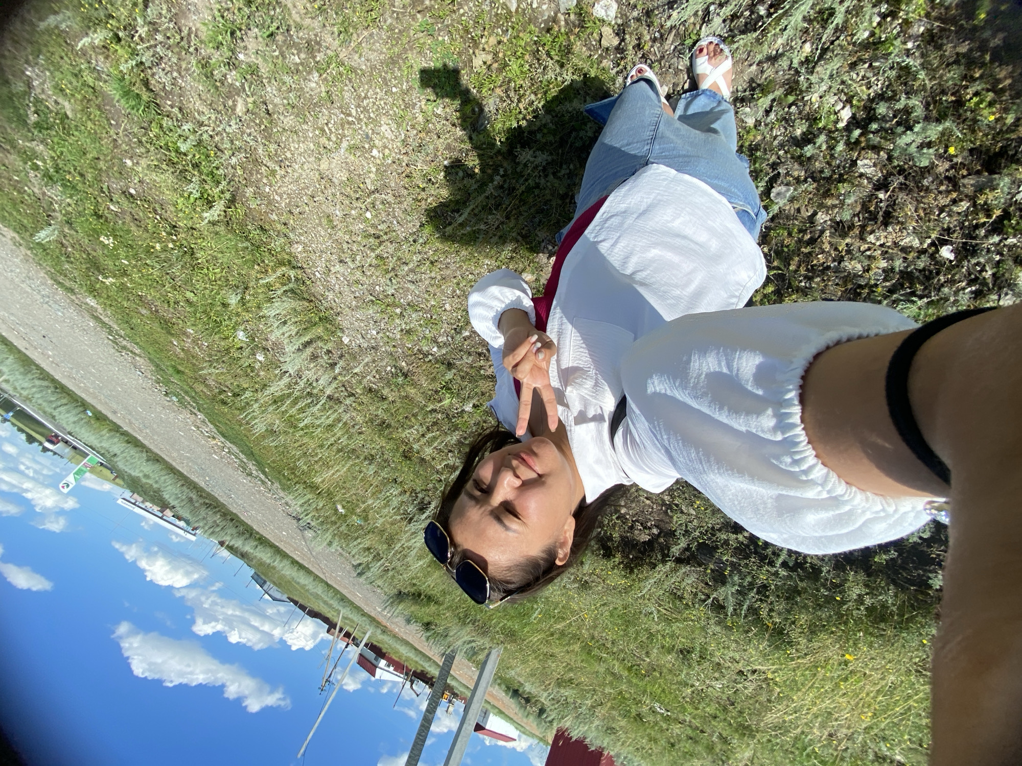 A young woman lies on her back in a grassy field, taking a selfie. She is wearing a white blouse, blue jeans, and white sandals. Her sunglasses are perched on her nose, and she is making a peace sign with her fingers. The field is dry and appears to be in a rural area, with a dirt road and some buildings visible in the background. The sky is blue with scattered white clouds, suggesting it is a sunny day. The overall mood is relaxed and playful. The woman's expression is cheerful, and she seems to be enjoying the outdoors. The image was taken in Yuldybayevo, Russia. There are no visible text elements.