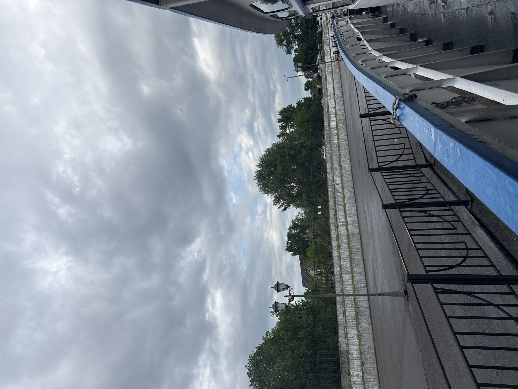 The image depicts a cloudy sky above a riverside promenade in Kazan, Russia. A promenade with a black metal fence runs along a water body, suggesting it is a river or canal. Beyond the fence, there is an asphalted area and a tiered concrete embankment. A vintage street lamp with two globes stands on the promenade. Lush green trees line the background, with glimpses of buildings and parked cars. In the foreground, the railing of a vessel, painted white with a blue stripe, is visible. The overall scene suggests a day with overcast weather, likely before or after rain, given the damp appearance of the asphalt.