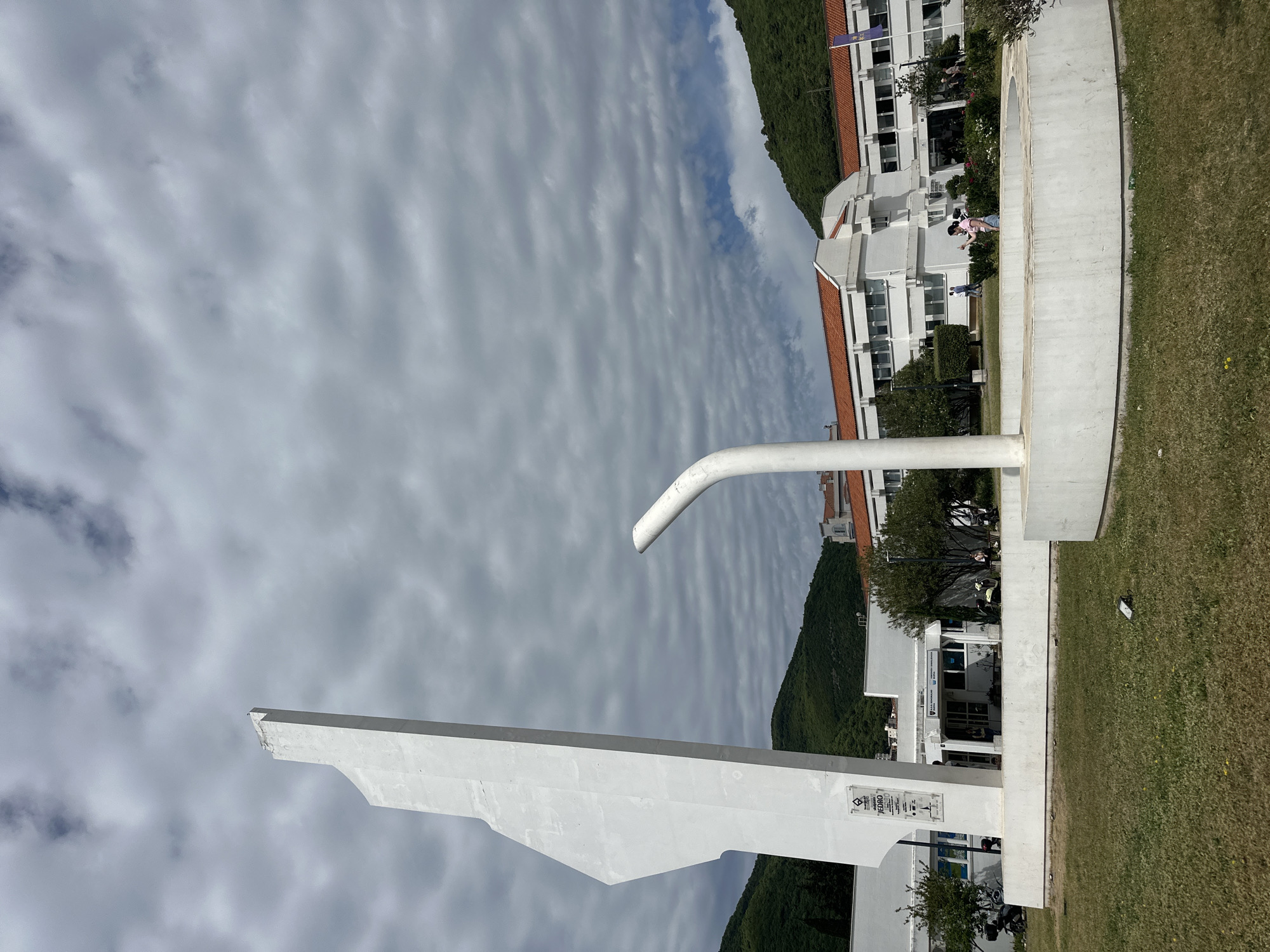 The image shows a white, abstract architectural sculpture set against a backdrop of a cloudy sky, buildings, and a green hillside. The sculpture consists of geometric shapes, including a long, flat, angled piece and a curved pipe-like element. The sculpture is situated on a concrete base.
In the background, there are white buildings with red-tiled roofs, typical of the Mediterranean architecture found in Montenegro. The hillside is covered in lush, green vegetation. People are walking near the buildings, appearing small in the context of the overall scene.
Based on the architecture, foliage, and general appearance, it is likely the image was taken in or around the city of Budva, Montenegro. Specifically, the sculpture is located near the main entrance to the Slovenska plaza complex (Hotel complex).