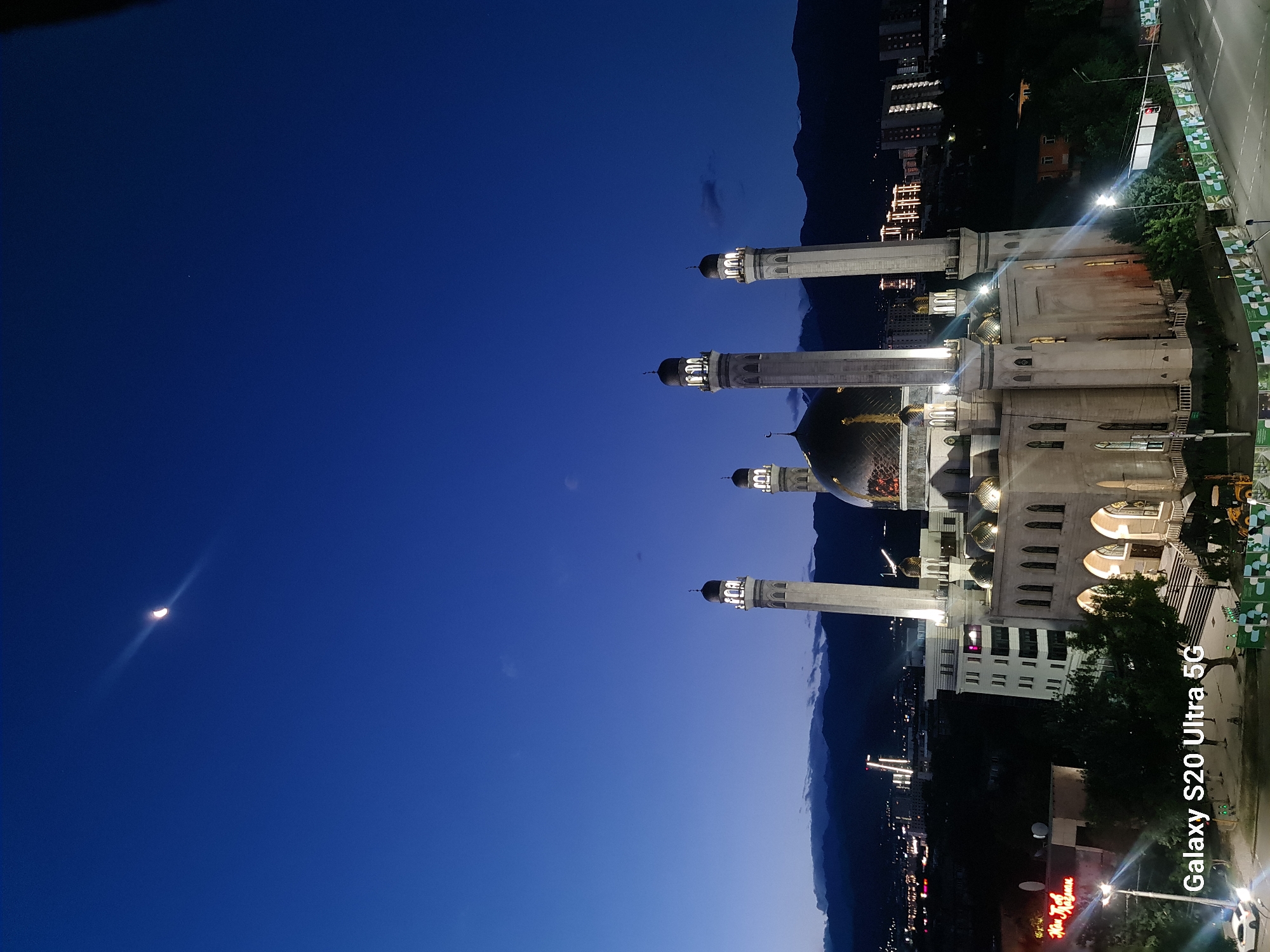 The image captures the Central State Mosque in Almaty, Kazakhstan, at dusk. The mosque, a prominent architectural landmark, is illuminated, highlighting its intricate design with multiple domes and minarets. The sky above is a deep blue, transitioning from twilight, with a crescent moon visible. In the background, the silhouette of the Zailiysky Alatau mountains provides a natural backdrop to the urban landscape. Buildings and city lights are scattered across the middle ground, indicating the presence of a bustling city. A white car is parked near the entrance of the mosque. The foreground features a street with traffic lights and some trees. The overall scene depicts a serene evening in Almaty, with the mosque standing majestically against the darkening sky and surrounding city.